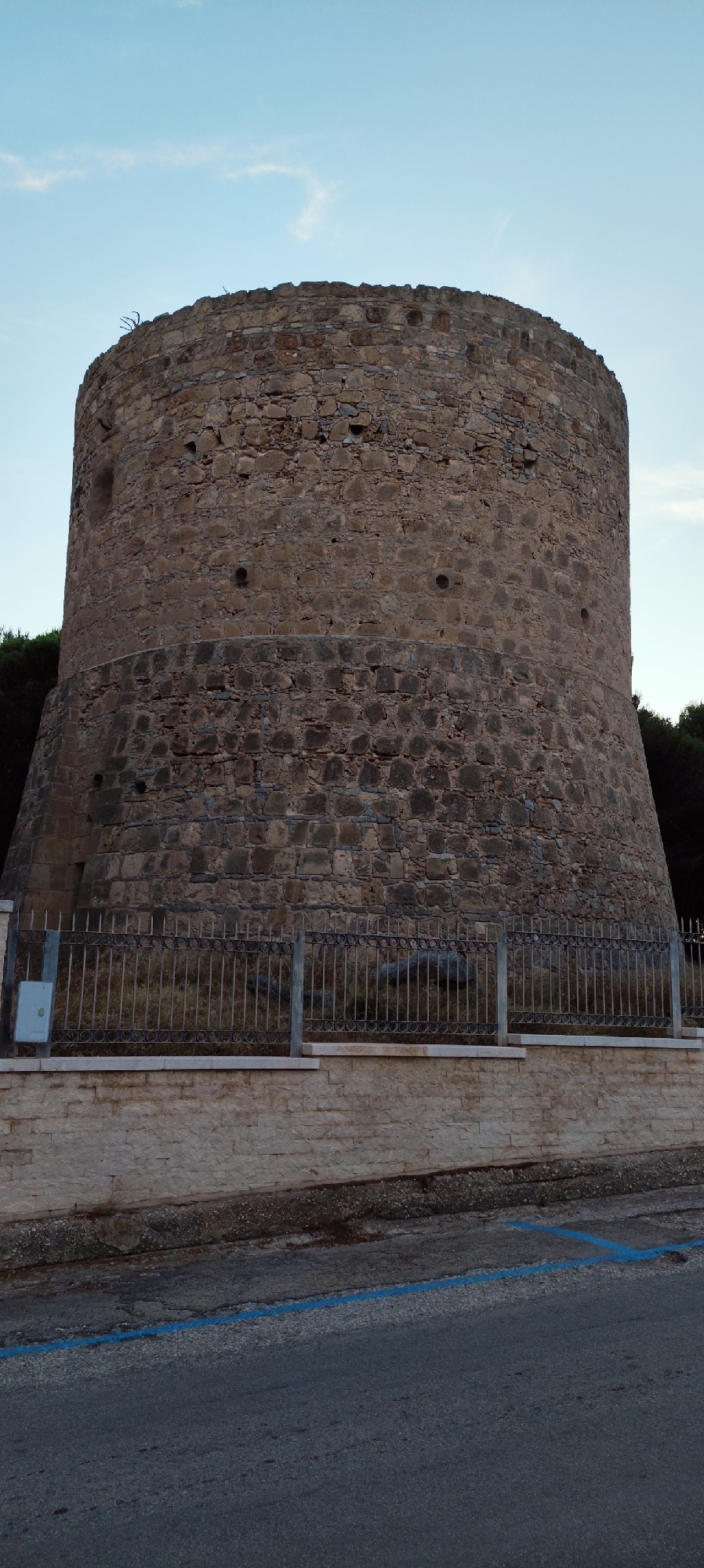The image shows a large, round stone tower, likely a historic structure, with a weathered appearance. The tower is constructed from irregular, light brown stones and has a slightly tapered cylindrical shape, wider at the base and narrowing towards the top. There are small, circular openings visible at various points on the tower's facade, potentially arrow slits or ventilation shafts. The upper rim of the tower appears uneven and shows signs of age.
The tower is situated in an outdoor environment. In front of the tower, there is a low wall constructed from lighter-colored, rectangular stones. A dark metal fence with a decorative circular pattern separates the area in front of the tower from the viewer's perspective. Behind the fence, the ground is covered with dry, yellowish grass and some scattered rocks.
To the left of the tower, a small, light-colored electrical box is mounted on a post. The background behind the tower is dominated by dark green foliage, likely trees, suggesting a natural setting. The sky above is a pale blue with a few wispy white clouds.
In the foreground, a dark asphalt surface is visible, with a bright blue painted line marking what appears to be a parking space or lane. The overall impression is of an ancient structure located in a semi-rural or park-like setting within or on the outskirts of a town. The specific location is the historic tower near the lighthouse in San Vito Lo Capo, Italy, a prominent landmark in the coastal town.
