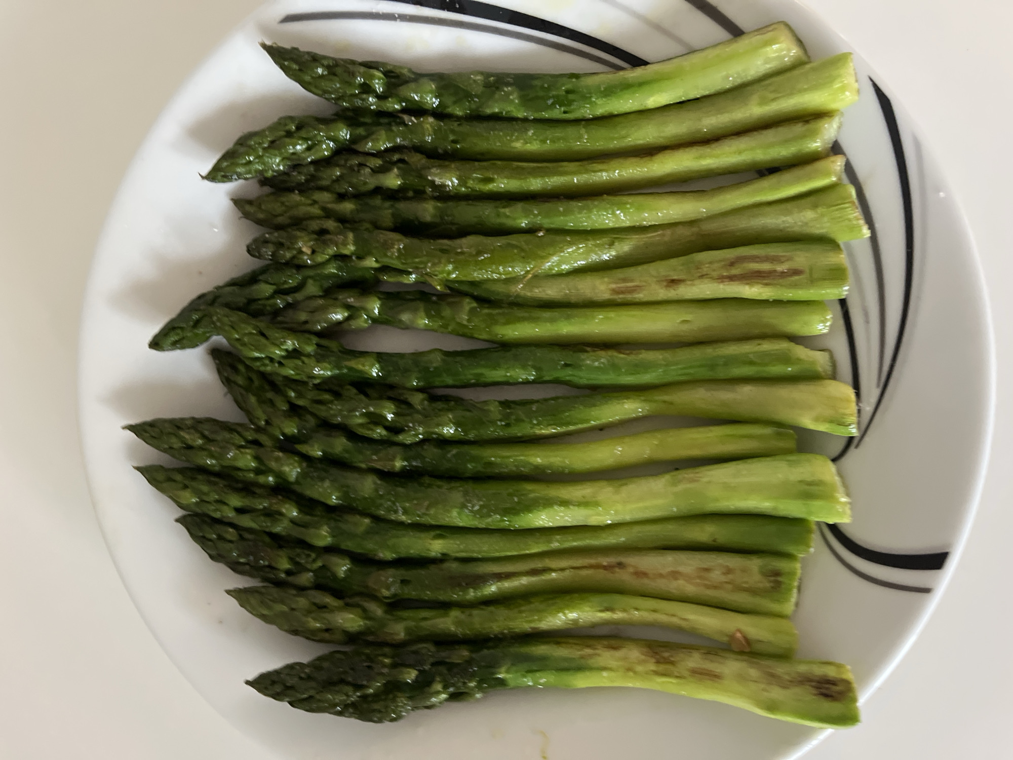 The image displays a generous serving of cooked asparagus spears neatly arranged on a white plate. The asparagus spears are laid out in rows, filling the majority of the plate's surface. They exhibit varying shades of green, from a darker, richer green at the tender tips to a lighter, more yellowish-green towards the stem ends. The spears appear glossy, suggesting they may have been lightly coated with oil or cooked with some moisture. Some sections of the asparagus, particularly towards the stem ends, show subtle brownish or slightly caramelized markings, indicative of roasting or pan-frying.
The plate itself is white with a distinctive pattern of black stripes, specifically two thicker lines and one thinner line, accentuating its rim on one side. The background is a plain, light-colored surface, likely a table or countertop, suggesting an indoor setting. The lighting is bright and even, casting minimal shadows and highlighting the texture and color of the asparagus. There are no people visible, nor is any specific activity taking place other than the plating of the food. No visible text is present in the image.