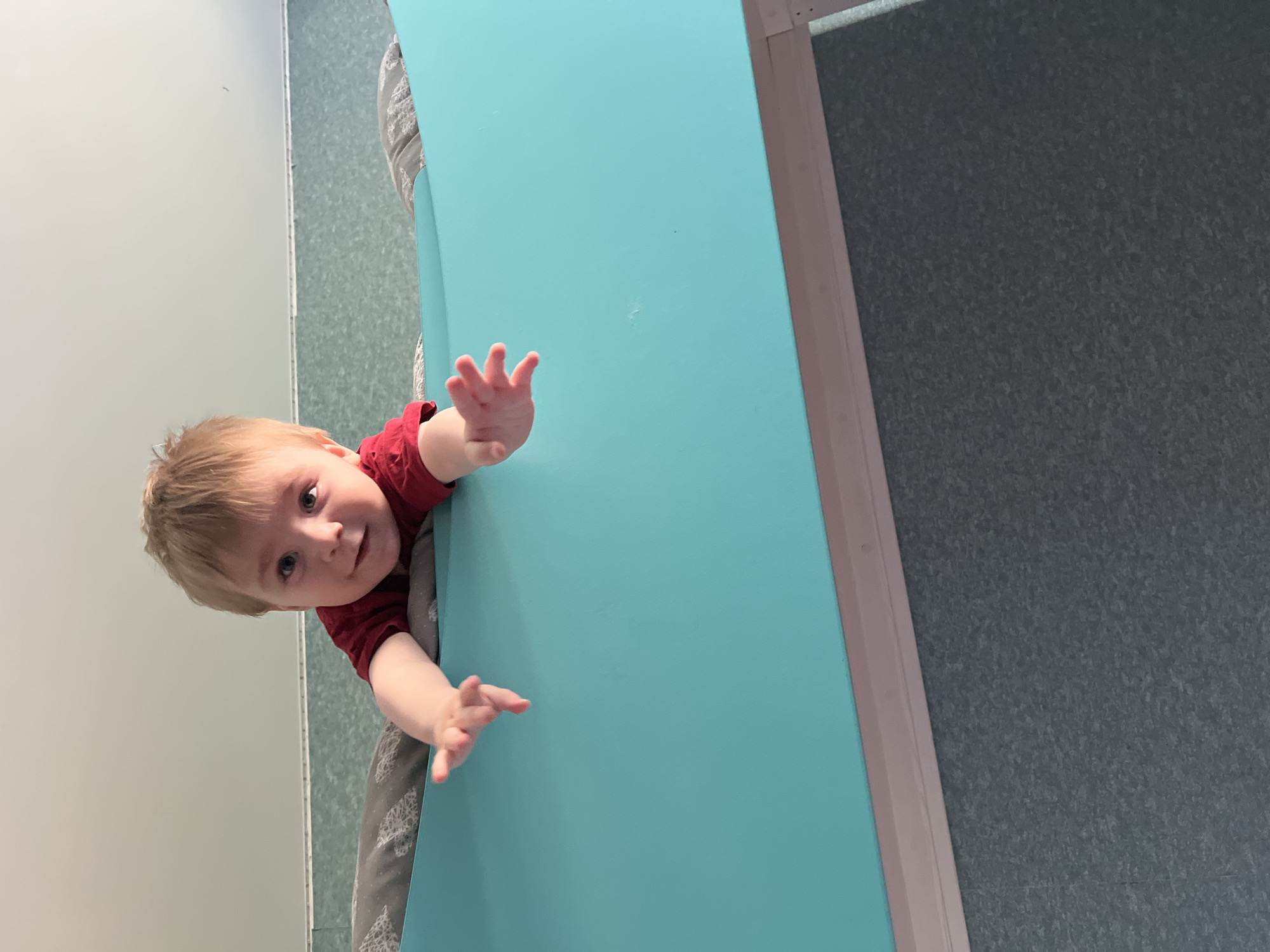 A young child with blond hair and a red shirt is leaning over a turquoise-colored changing table. The child's hands are reaching out towards the viewer. The changing table appears to be in a public restroom or similar facility, as indicated by the gray textured wall and silver trim. The location of the scene in the city of Noviy, Russia is not discernible from the image.