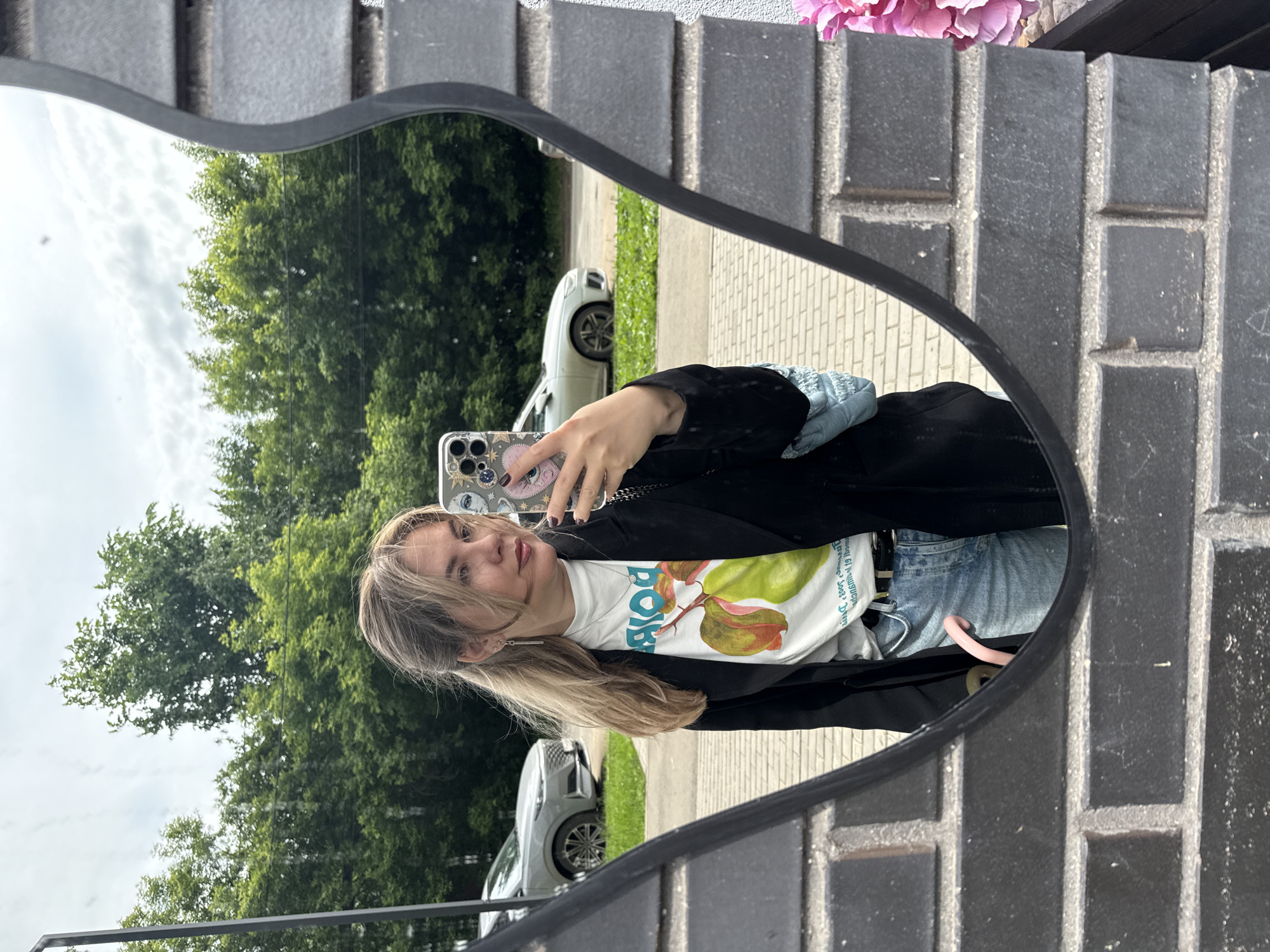 A young woman is captured in a mirror selfie, holding her smartphone, which is adorned with a patterned case featuring eye designs. She has shoulder-length, light brown hair and is looking towards her phone screen with a neutral expression. She wears a black blazer over a white t-shirt that displays the word "DOPE" in green letters, along with two stylized pears and partially visible Cyrillic text, likely "С Днем рожденья!" (Happy Birthday!). She also wears blue denim jeans, from which a pink object, possibly a hair tie, hangs. A light blue, possibly puffy-sleeved top or bag, is visible on her left side. Her nails are painted dark.
The selfie is taken in a unique black-framed mirror with an irregular, wavy top edge. The reflection in the mirror reveals an outdoor scene under a cloudy daytime sky. Lush green trees, a paved walkway, and two parked cars (one white, one grey/silver) are visible in the background. Outside the mirror's frame, the real-world setting consists of a dark grey/black brick wall with lighter mortar, and some pink artificial flowers are visible in the upper right corner. The overall setting is outdoors, possibly in a street or residential area in Izhevsk, Russia.