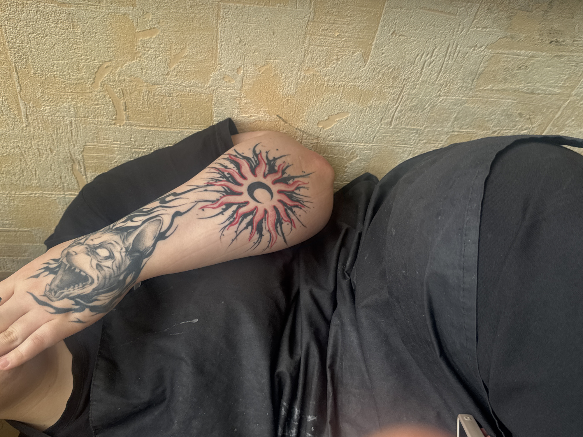 The image shows a close-up of a person's arm with tattoos. On the upper part of the arm and hand, there is a black and grey tattoo of a wolf's head with flames. Further down the arm, on the elbow area, there is a tattoo of a sun with a crescent moon in the center, rendered in black and red. The person is wearing a black apron or shirt. The background is a textured, light-colored wall. The image is taken indoors, with no visible indication of the city of Zaokskiy, Russia.