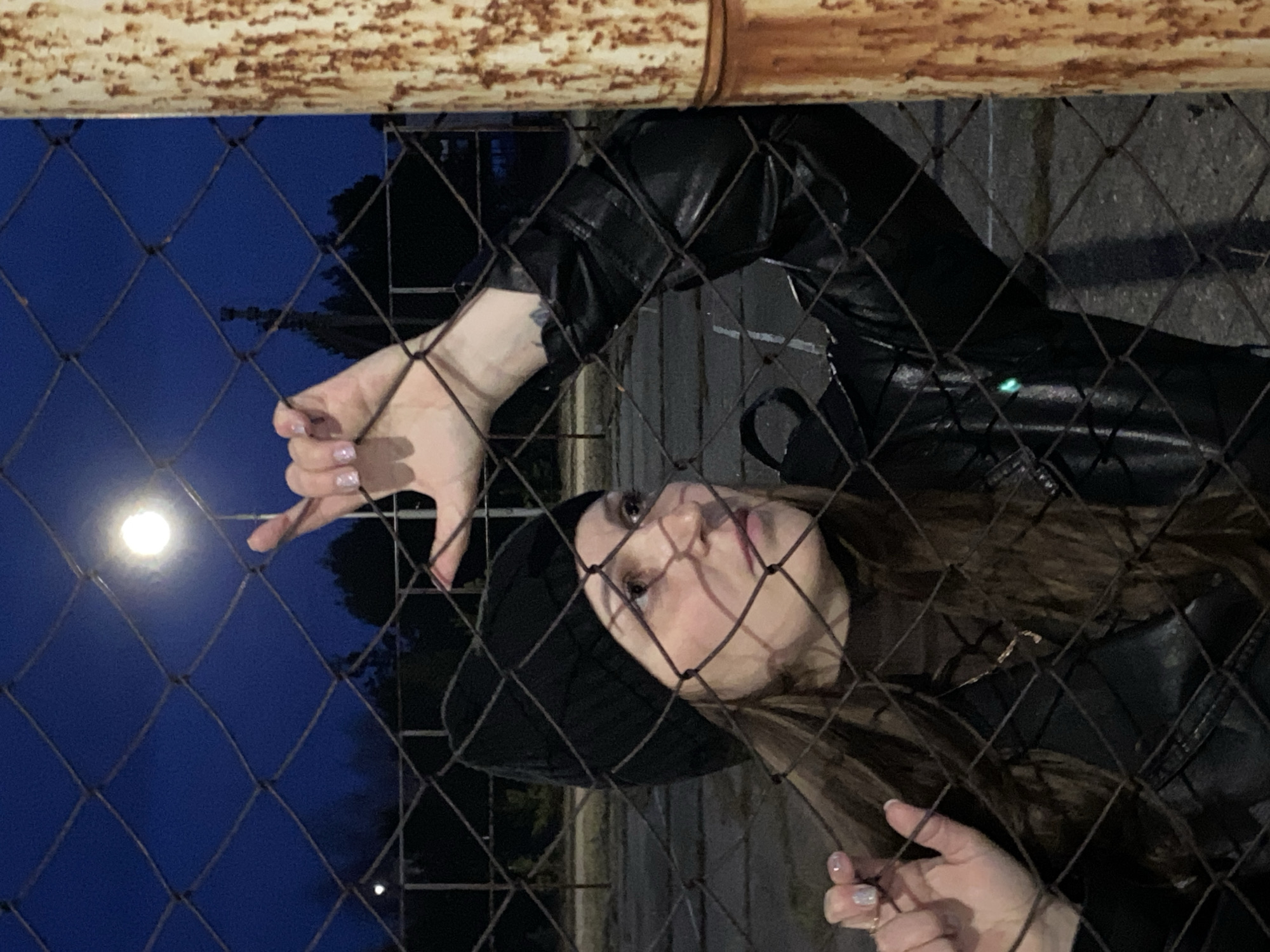 A woman is seen through a chain-link fence with a moon visible in the background. The location is presumably somewhere in Марковский, Russia. The woman is wearing a black leather jacket and a dark hat. She is looking up and towards the moon, with her hand held up in front of it. A rusted metal bar is visible at the top of the frame, likely part of the fence structure. The background is dark, suggesting it is nighttime.