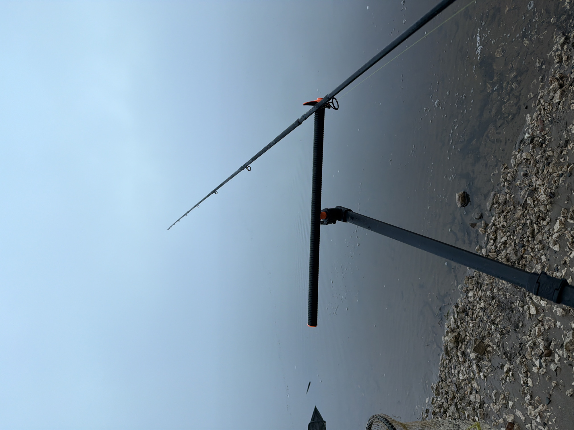 The image, rotated 90 degrees clockwise, displays a peaceful fishing scene under an overcast sky. A black fishing rod, equipped with guides and a fishing line, extends from the lower right towards the upper left. It is held steady by a dark, ribbed rod holder or bank stick, which features subtle orange accents and is planted firmly into the ground or bank. The fishing line disappears into the calm, dark water.
The upper portion of the frame shows a uniformly light blue-grey, cloud-covered sky, indicating an overcast day. The water beneath the fishing rod is still and murky, reflecting the sky. Scattered small bubbles and faint ripples are visible on the water's surface, and tiny water droplets adhere to the fishing rod, suggesting humidity or recent rain.
Along the right side and extending into the lower frame, a rocky or pebbly shoreline is visible. It consists of numerous light-colored stones mixed with darker sand or sediment. Partially visible at the bottom of the frame, near the shore, is a segment of a fishing net, possibly a landing net or a keepnet. A small, dark, elongated object, likely a small fish or debris, is in the water near the rod holder. No people or discernible text are present in the photograph. The scene depicts a quiet moment of fishing by a body of water, consistent with a riverbank or lakeside environment in Chaikovsky, Russia.