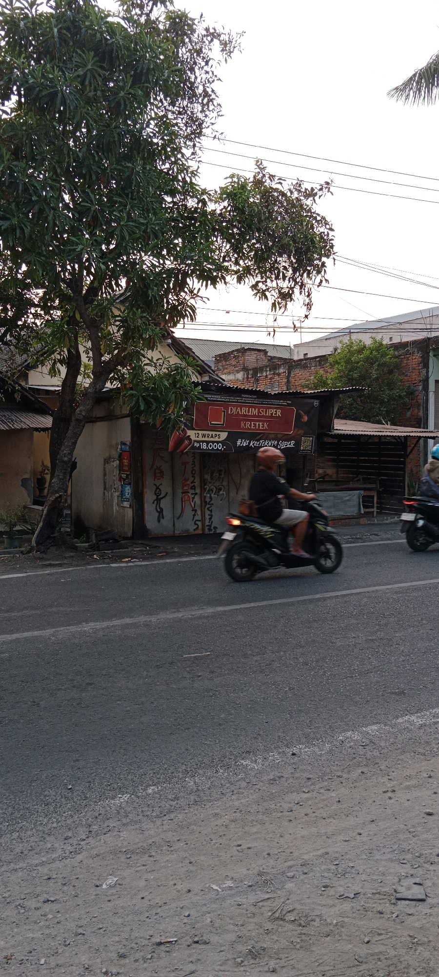 The image depicts a street scene in Bojonegoro, Indonesia, characterized by a tree with dense foliage on the left, partially obscuring a building with a weathered facade covered in graffiti and signage. A prominent banner for "DJARUM SUPER KRETEK" is visible, advertising "12 WRAPS Rp 18.000,-" and a hashtag "#JIWI KRETEKNYA SUPER". Two individuals on motorcycles are captured in motion. The rider in the foreground, wearing a dark shirt, light shorts, and an orange helmet, is blurred due to their movement. Their motorcycle is dark and appears to have yellow and green decals. To the right, another motorcycle and rider, partially visible and also blurred, are traveling in the same direction. The street itself is asphalt, with a white line marking the lane. The background shows the roofs of buildings, including some with corrugated metal and brick structures, under an overcast sky. The overall impression is a typical urban street environment in Indonesia, with elements of commerce, local life, and traffic.