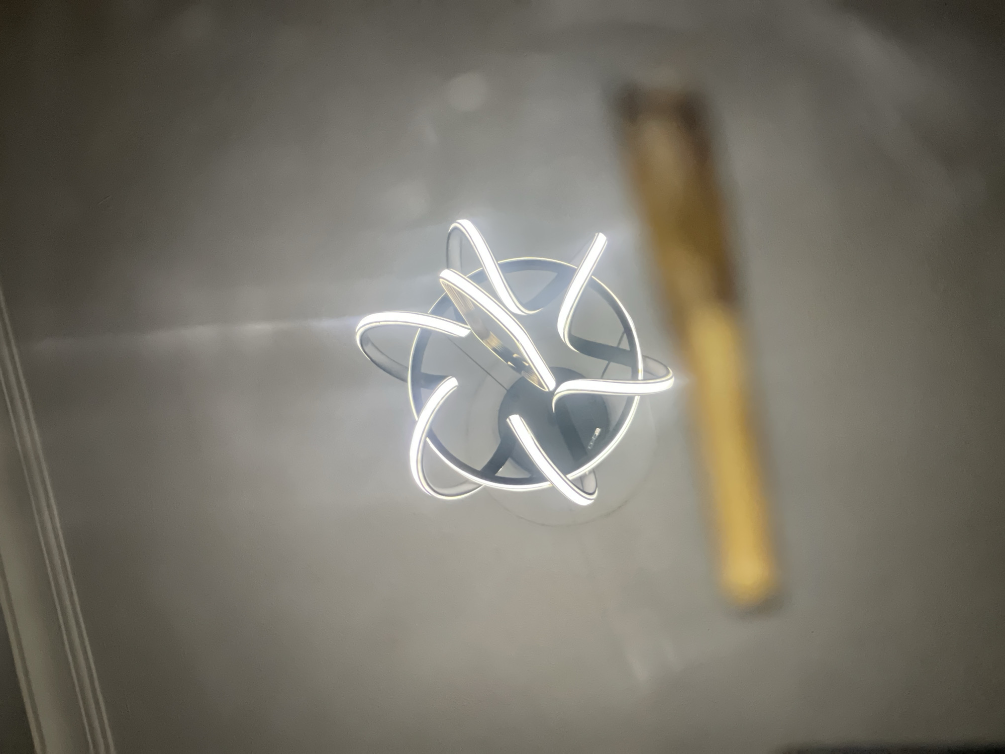 A ceiling-mounted, modern light fixture is the focal point, featuring a multi-layered, stylized design with illuminated white bands against a dark, circular background. Out of focus and in the background to the right is an object that appears to be a golden pole or tube. The environment appears to be an indoor space, likely a room within a building, given the presence of the ceiling. No people are visible in the image. The location in Abuja, Nigeria cannot be determined from the visual information provided.