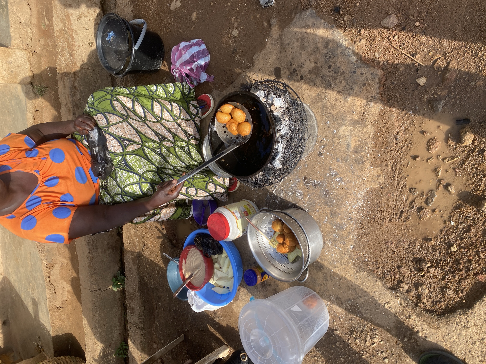 In Jos, Nigeria, a woman is seen preparing food outdoors, likely a street food vendor. She is wearing an orange, blue-dotted t-shirt and a green and white patterned skirt. In front of her, a large black pot sits on a metal stand filled with burning coals, where oil is being heated for frying. A slotted spoon is used to remove golden-brown fried fritters from the hot oil, and these are placed in a metal colander. Nearby, other food preparation items are visible: a blue basin containing white food items, a red bowl with a thick, light brown mixture, a large jar with a red lid, a small blue container, and a clear plastic container. A black bucket with a handle is also present. The ground is a mixture of dirt and concrete, with puddles of water visible, suggesting recent rain or water spillage. The scene is illuminated by natural sunlight, casting distinct shadows.