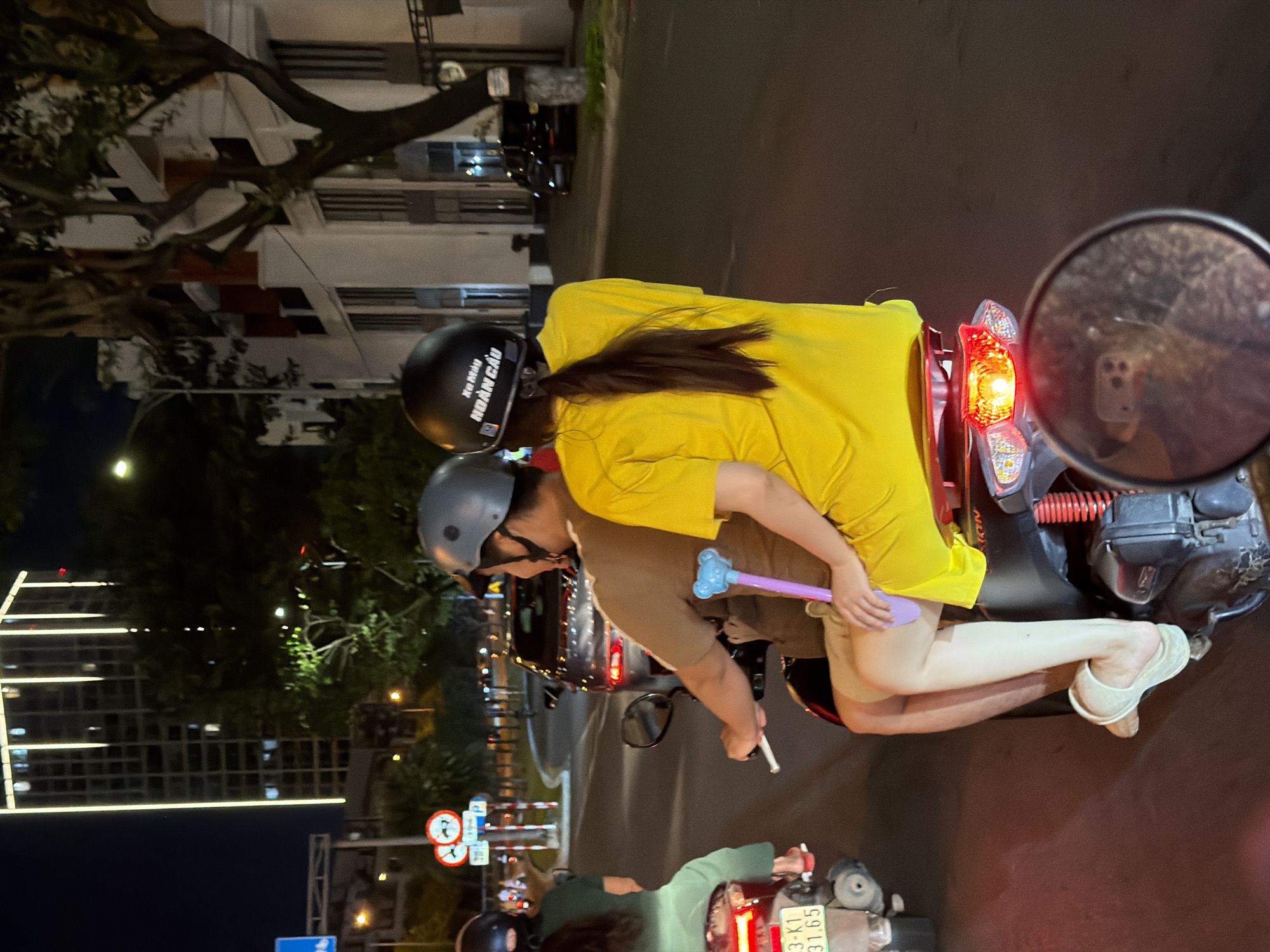 The image captures a nighttime scene on a street in Da Nang, Vietnam, likely a busy thoroughfare given the presence of multiple vehicles and illuminated buildings. In the foreground, two people are riding a scooter. The rider, wearing a gray helmet, is looking to the right. The passenger, a woman with a black helmet that has "Xe Máy HOÀNG CÂU" written on it, is facing forward. She is wearing a bright yellow t-shirt dress and has a purple object with a blue bear on it tucked under her arm. Her long dark hair is tied in a ponytail. The scooter's taillight is illuminated, and a portion of a side mirror reflects a smartphone with a cracked screen and a blurry image on it.
To the left of the scooter, another person on a scooter is visible, wearing a green shirt and a helmet. Their scooter has a license plate that reads "3-K1 31.65". In the background, a dark-colored car is stopped at an intersection. Traffic lights and street signs are visible, indicating an active urban environment. Further in the background, a tall, modern building with vertical lights is illuminated against the night sky, and other buildings line the street, some with visible architectural details and lighting. Trees with dark foliage are also present, contributing to the urban landscaping. The overall impression is one of everyday urban life in Da Nang at night.
