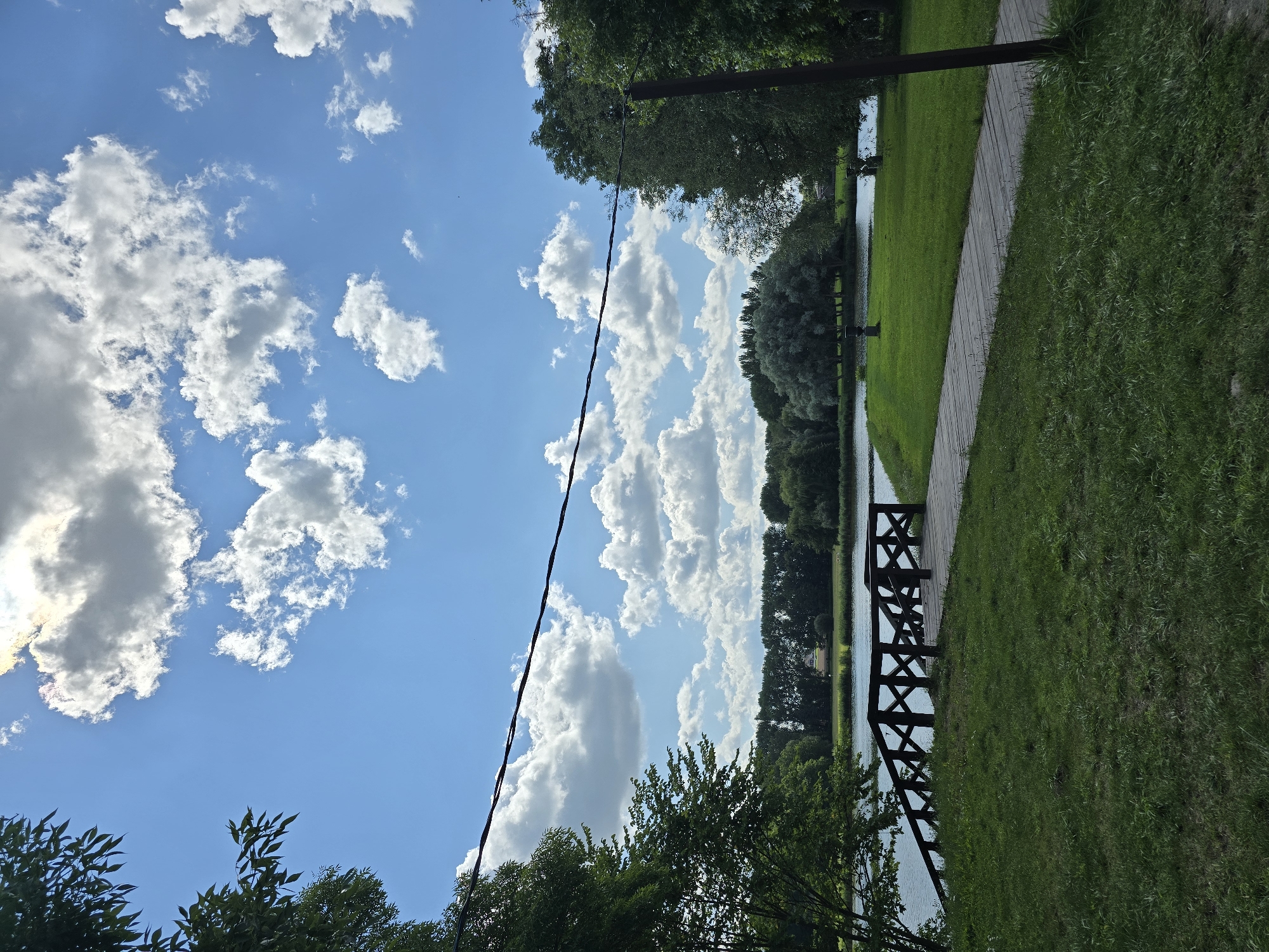 The image depicts a serene park landscape in Niasvizh, Belarus. A wooden walkway curves through a grassy area towards a body of water. To the left, a wooden bridge with a lattice railing extends over the water. Lush green trees line the far bank of the water, with a treeline and some buildings visible in the distance. The sky above is a vibrant blue, dotted with fluffy white clouds. A dark wire is strung across the upper portion of the image. The scene is bathed in daylight, suggesting a pleasant outdoor setting.