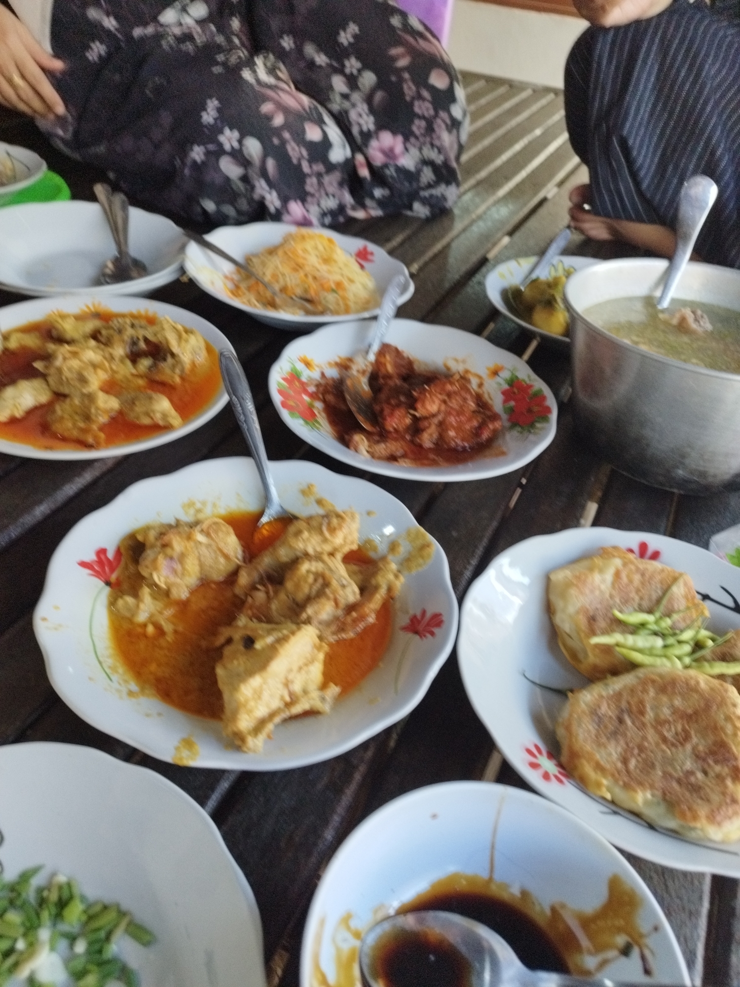 In Petapan, Indonesia, a table laden with food is displayed. The table appears to be made of wood. Several plates of food are arranged on the table. One plate contains what looks like fried chicken in a yellow sauce, another contains a reddish meat dish, and a third has a portion of noodles. There is also a pot of soup with meat and vegetables visible. Two individuals are partially visible, seated at the table and appear to be eating. The image is taken from a top-down perspective.