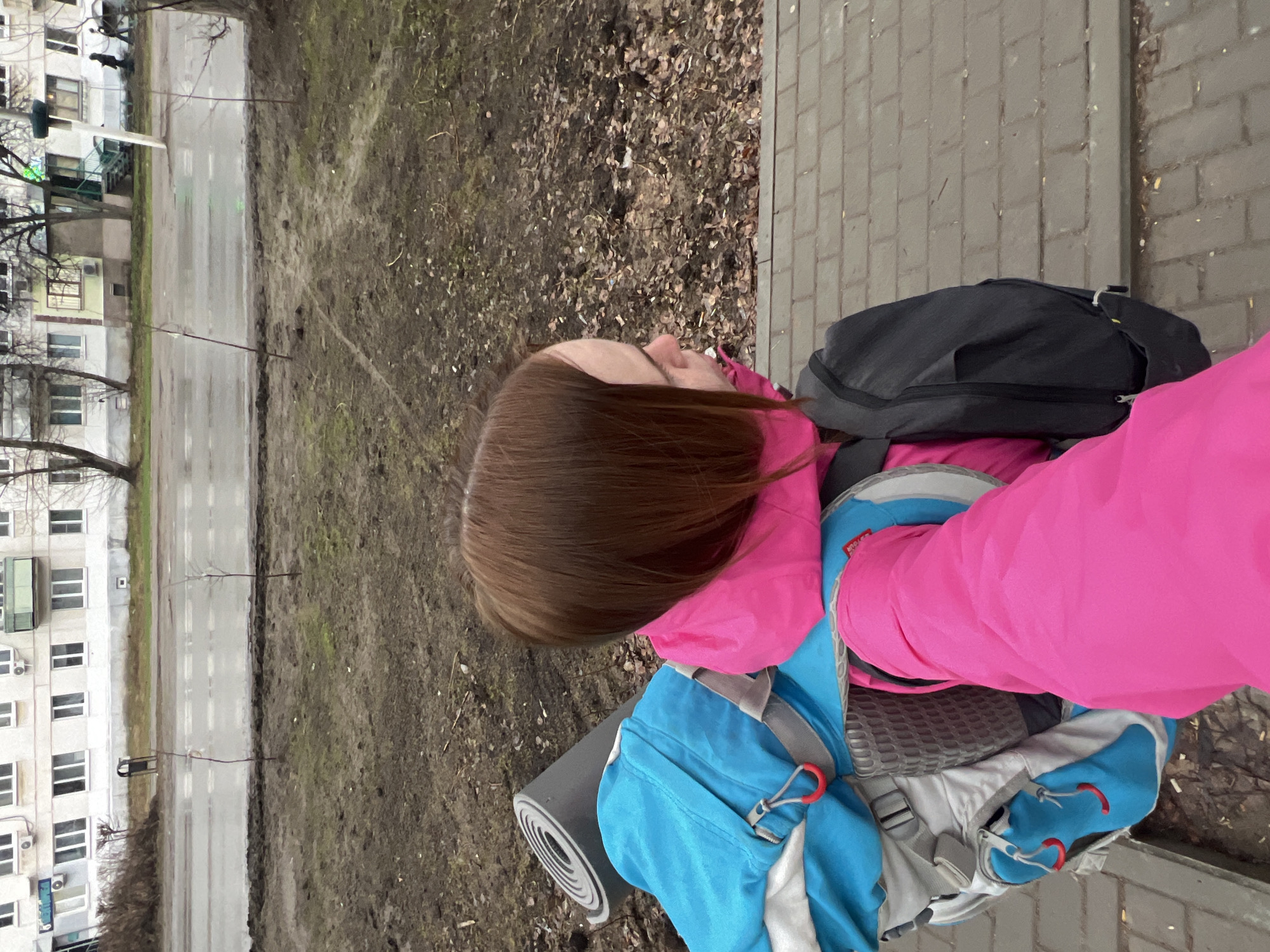 A person with brown hair, wearing a pink jacket and a blue backpack with a sleeping mat attached, stands on a brick sidewalk. To the left, there is a street and a multi-story building with white walls and many windows, typical of urban residential architecture. A dirt patch is visible between the sidewalk and the street. Given the information provided, this is likely a scene in Chaikovsky, Russia.