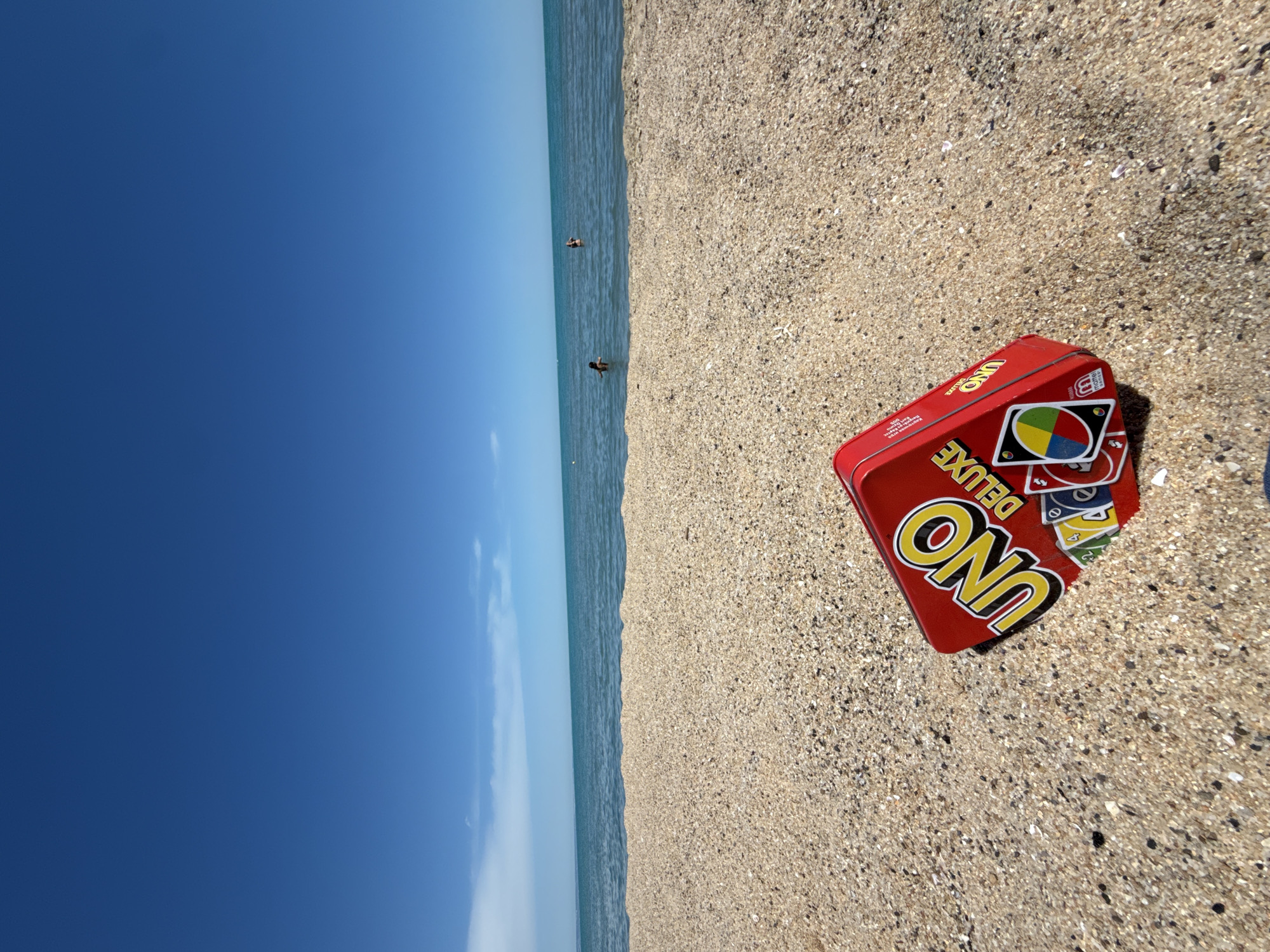 The image depicts a bright, sunny day at a sandy beach. The foreground is dominated by a red tin of UNO Deluxe cards partially buried in the sand. The cards themselves, with their colorful faces visible, are also partly obscured. Beyond the beach, a calm, blue sea stretches to the horizon, meeting a clear, vibrant blue sky with a few wispy white clouds. In the distance, two individuals can be seen wading in the shallow water. The overall scene evokes a sense of leisure and outdoor recreation. The location is not identifiable as Varna, Bulgaria, or any other specific city.