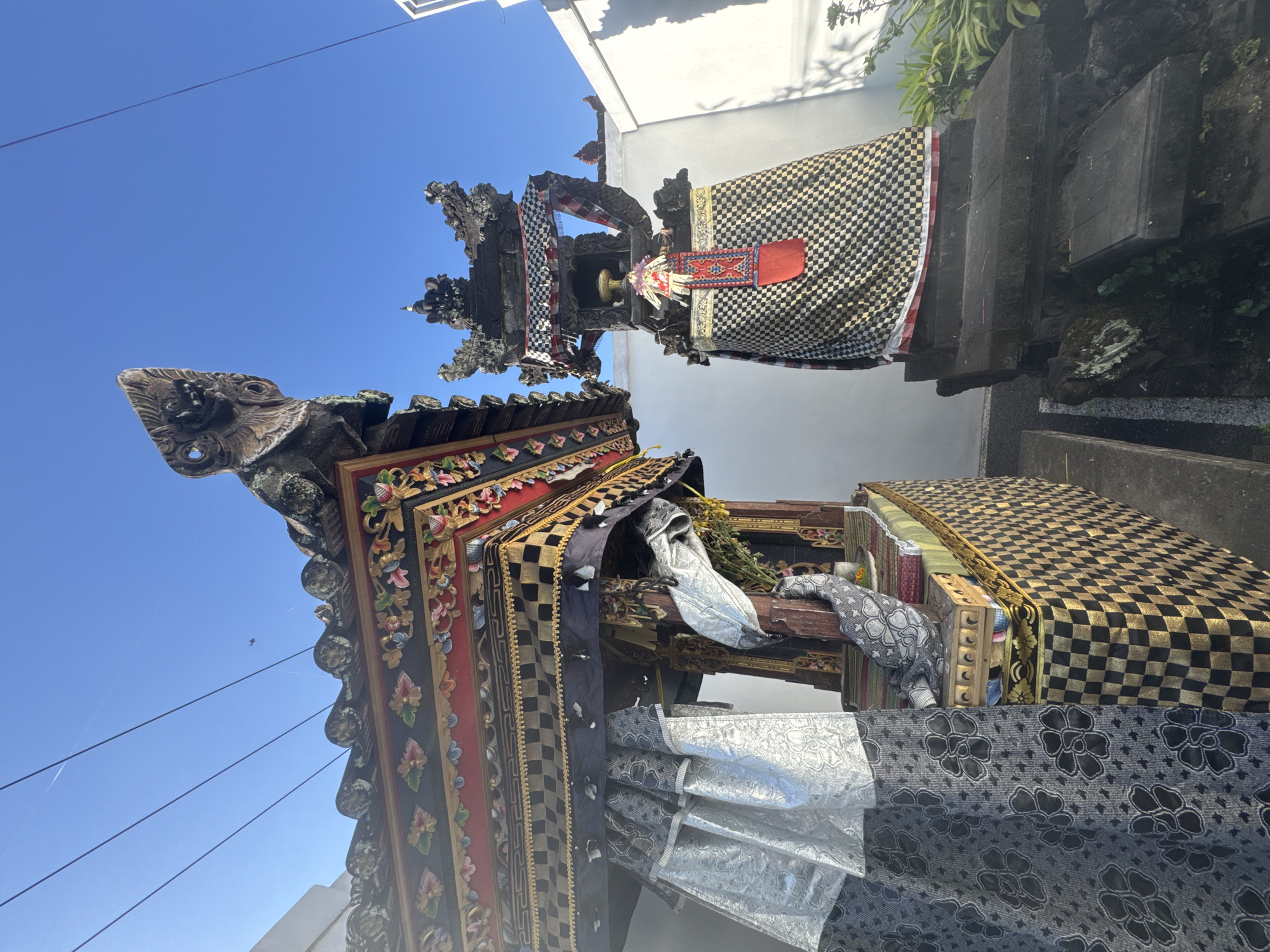 The image depicts two ornate Balinese shrines in what appears to be an outdoor setting in Mengwi, Indonesia. The shrines are elaborately decorated with intricate carvings, checkered patterns, and fabric adornments. The shrine on the left is larger and features a multi-tiered roof with colorful floral motifs and what looks like a carved mythical creature at the peak. It is draped with flowing silver and black patterned fabrics. The shrine on the right is also richly decorated with a similar checkered pattern and a tiered, pagoda-like roof topped with decorative elements. Both shrines are adorned with ceremonial cloths, including red and white checkered fabric. The background consists of a bright blue sky and a white wall, suggesting a sunny day. The shrines are positioned on stone bases, and some greenery is visible near the base of the right shrine. The overall scene conveys a sense of cultural and religious significance.