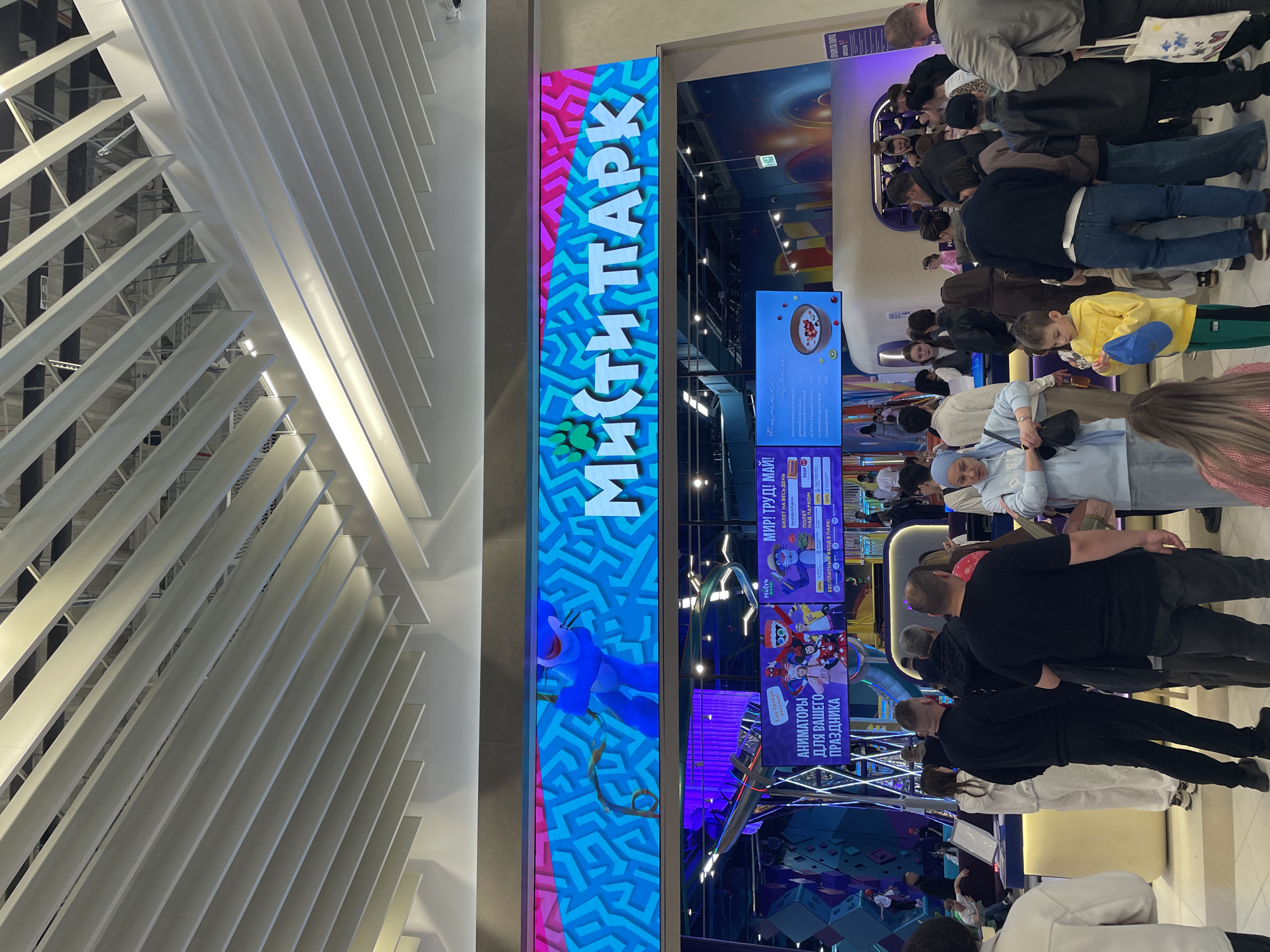 The image depicts the entrance to "Misti Park," an entertainment venue in Kazan, Russia, as indicated by the signage. A crowd of people is gathered at the entrance, presumably waiting to enter or purchase tickets. The signage includes the park's name and information on pricing and services such as “Animators for your holiday.” The architecture features a modern, angular ceiling design with white structural elements. The scene is indoors and well-lit.