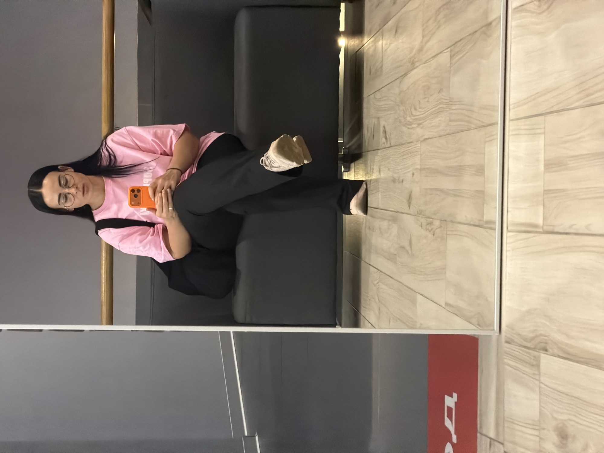 A young woman with dark hair and glasses is sitting on a gray sofa. She is wearing a pink t-shirt and black pants. She is holding a bright orange smartphone. Her legs are crossed, and she is wearing a sneaker on one foot and a sock on the other. The sofa is placed against a gray wall, next to a wood-look tiled floor. The photo appears to be taken inside a building in Saint Petersburg, Russia, as evidenced by the red-and-gray promotional signage partially visible near the bottom of the frame. The image suggests a rest or waiting area, likely in a public space. The photo is oriented vertically, as if taken from a mirror or reflective surface.