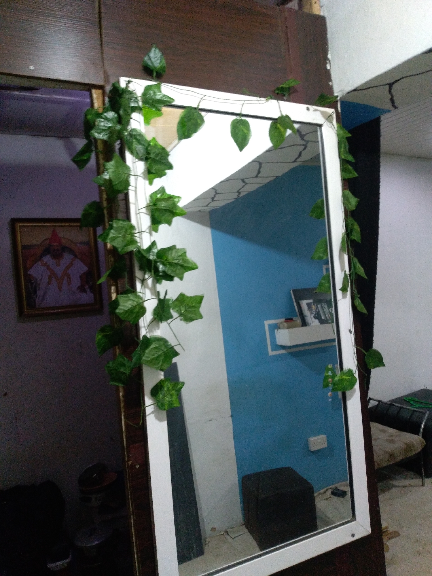 The image shows a full-length mirror with artificial ivy vines draped around its frame. The mirror is positioned in a room with a dark wooden cabinet on the left and a blue wall on the right. In the reflection, a white shelf with some items is visible on the blue wall. A framed picture of a person in traditional attire hangs on the wall to the left of the mirror. The floor appears to be tiled or concrete. There are no people visible in the image. The scene is likely a domestic interior.