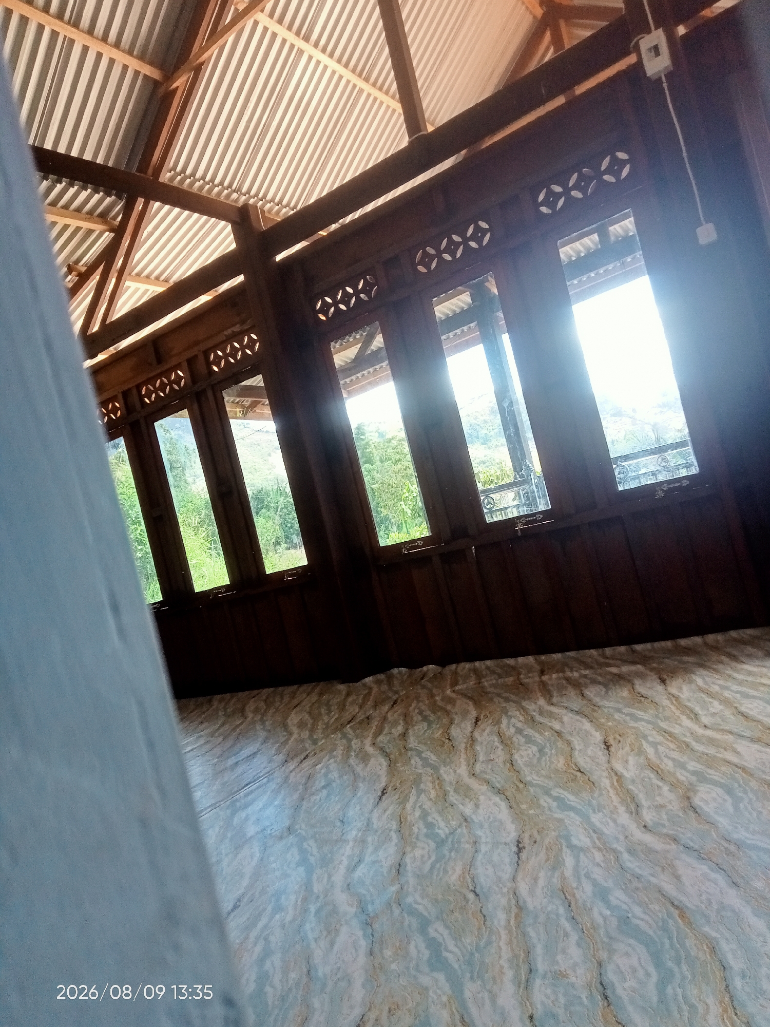 The image shows the interior of a simple, open-air structure with a corrugated metal roof supported by wooden beams. The walls are made of dark wood, and several tall windows with decorative vents above them look out onto a lush green landscape. The floor has a marbled pattern. A light blue fabric is partially visible in the foreground on the left. The timestamp "2026/08/09 13:35" is overlaid on the bottom left corner of the image. The scene is likely in the city of Perangian, Indonesia.