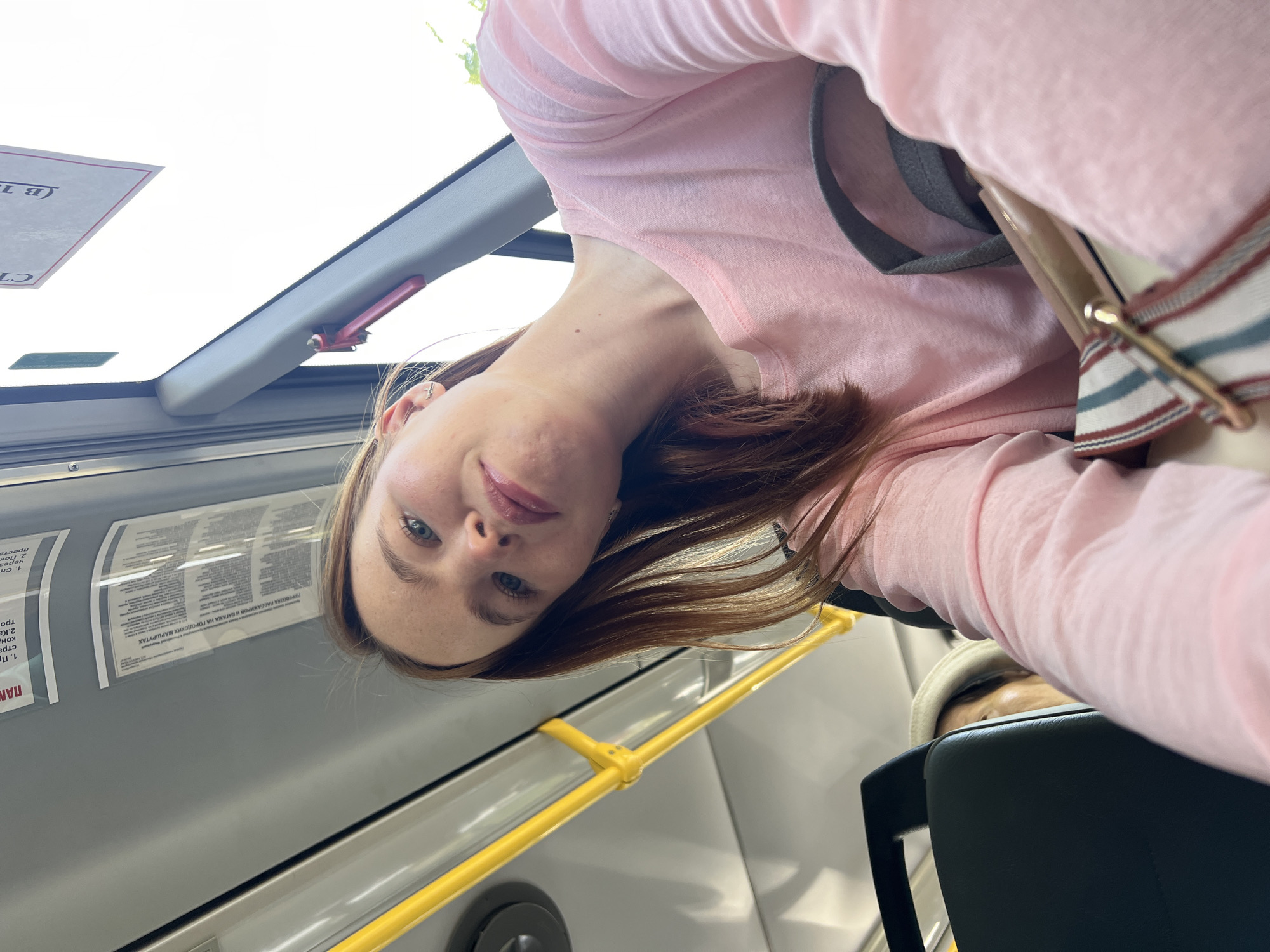 A young woman with fair skin and light brown hair is in a bus in the city of Chaikovsky, Russia. She is wearing a pink long-sleeved shirt and has a grey and white backpack. The interior of the bus is visible with yellow handrails and grey walls. Notices in Russian are posted near the window.