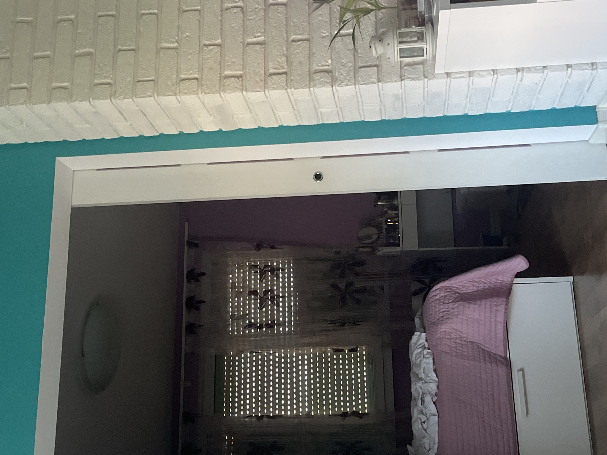 This is an indoor shot of what appears to be a doorway to a bedroom. The doorway has a white frame and a light blue wall on the left. The bedroom has a window covered by a sheer curtain with a floral design. A bed with a pink quilt and white ruffled sheets is partially visible. A white wardrobe is standing next to the bed. A light fixture is visible on the wall outside of the bedroom on the left side of the doorway. The room is mostly lit by the sun shining through the window. The location is potentially a building in Pokrovnik, Bulgaria.