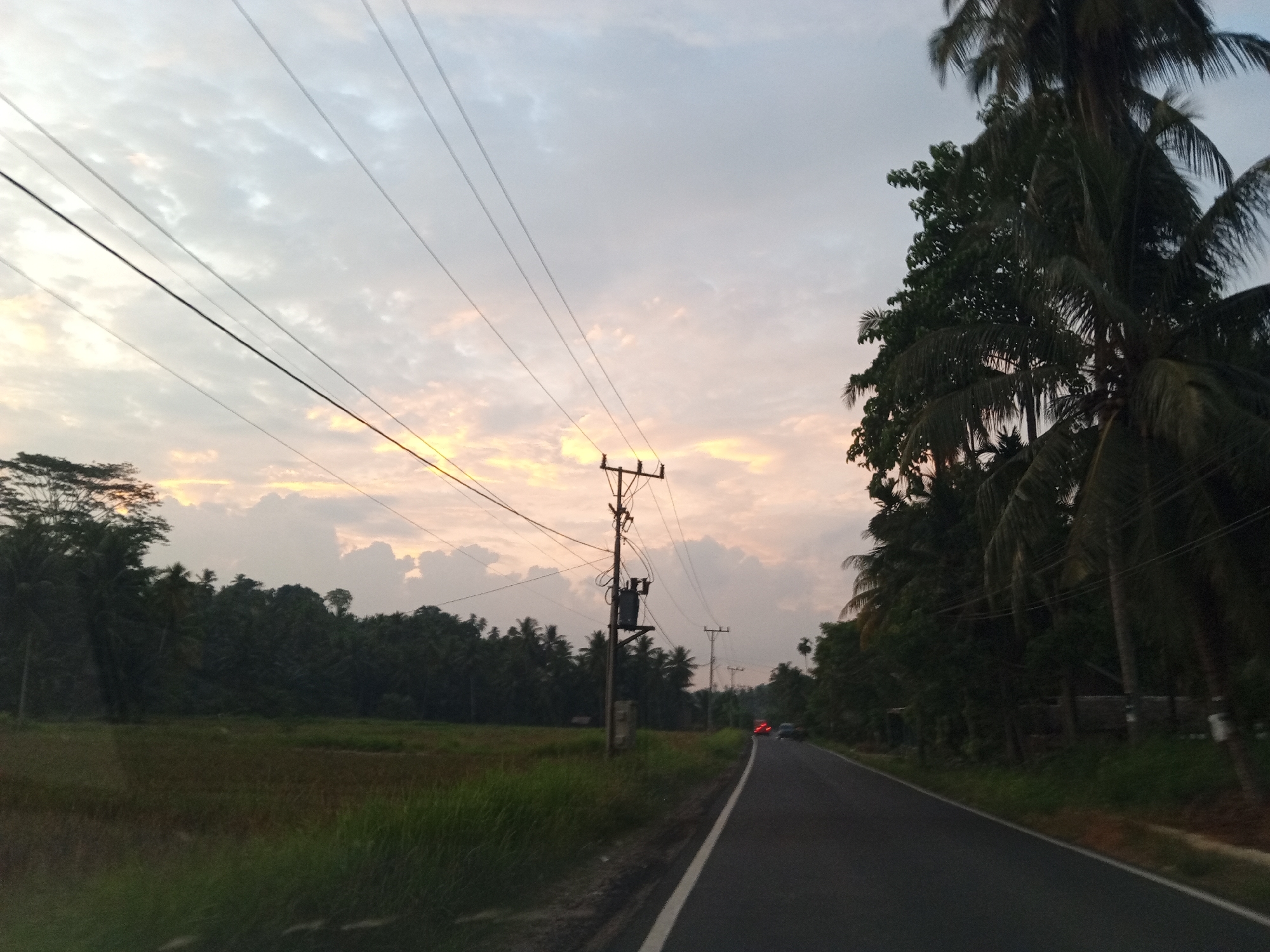 A scenic view of a rural road in Luengbaro, Indonesia, captures the tranquil beauty of the countryside at dusk. The sky is adorned with a soft palette of pastel colors, with hints of orange and pink illuminating the clouds. A line of lush green trees, including tall palm trees, borders the right side of the road, adding a tropical feel to the scene. On the left, a vast field of grass stretches out, with a dense forest forming the backdrop.
A power pole stands tall, its wires extending across the sky, connecting to other poles in the distance. A transformer box is mounted on the pole, indicating the presence of electricity in this remote area. A road, asphalted and marked with a white line, curves into the distance, inviting travelers to explore the picturesque landscape. In the distance, two cars with their headlights on are visible, hinting at the movement of life in this serene setting. The overall atmosphere is one of peace and tranquility, a perfect escape from the hustle and bustle of city life.