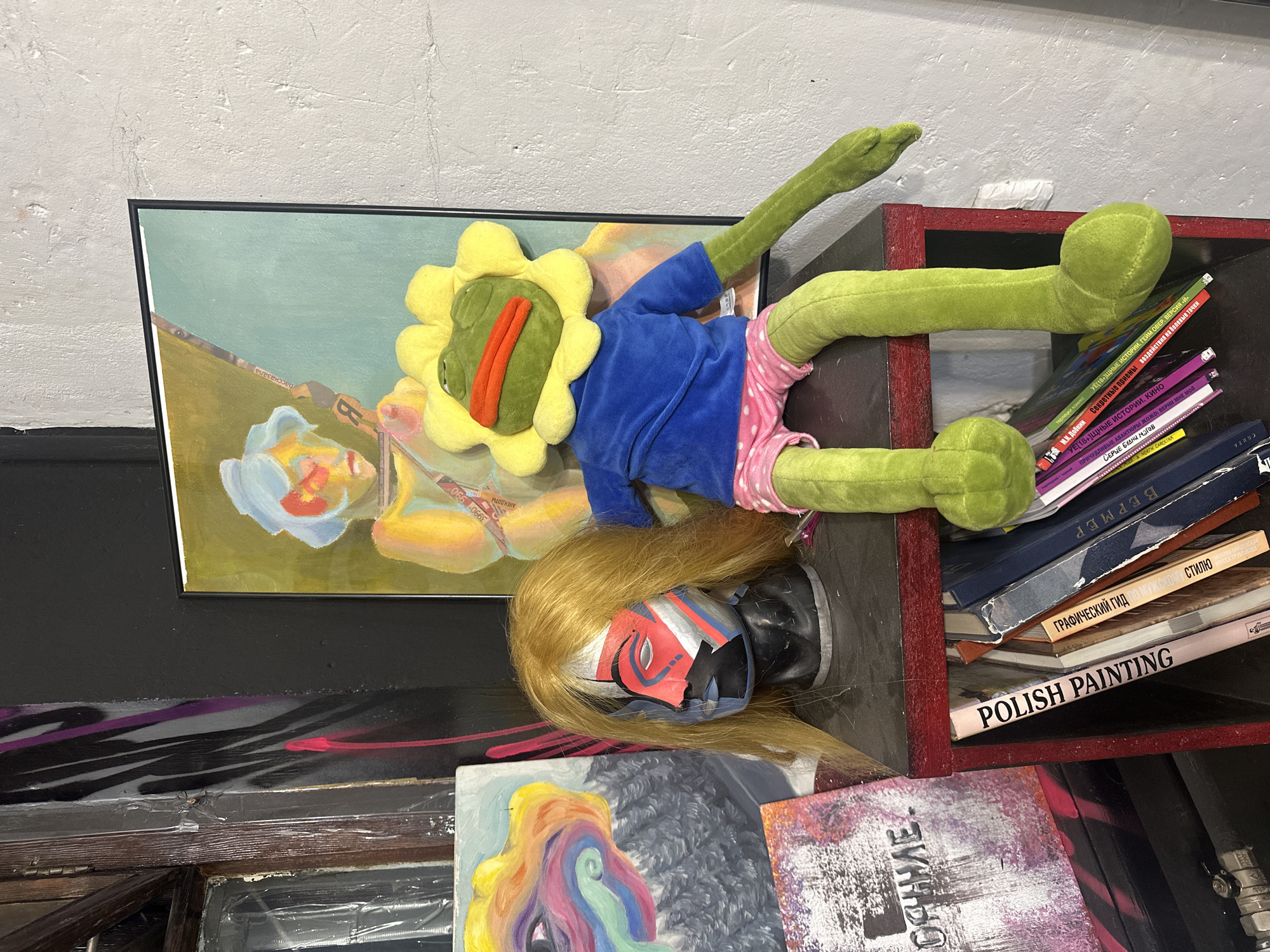 The image shows a cluttered space with various objects. On a shelf or surface against a gray wall, there is a framed painting depicting figures in an impressionistic style. A plush toy resembling a flower-headed figure with a blue shirt and pink shorts lies on top of the painting and next to a stack of books. A doll head with blonde hair and a face covering is positioned below the toy. The books are titled in Russian and English, with "POLISH PAINTING" clearly visible. The overall scene is somewhat chaotic with multiple art pieces and objects stacked together. The location is Moscow, Russia, based on the Russian text on the books.
