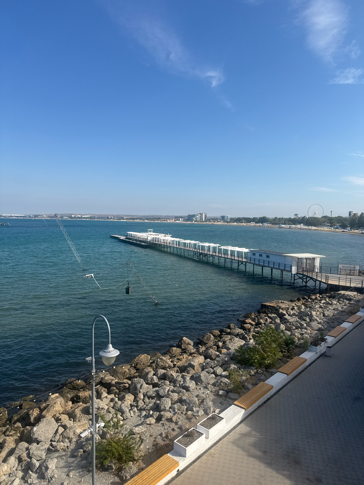 An aerial view of the Anapa embankment, located in Anapa, Russia, shows a white pier extending into the turquoise sea. The pier has a covered section at its end and is supported by numerous pilings. To the left of the pier, a water-ski cable system is visible, with poles and cables stretching across the water. The shoreline is lined with a rocky embankment and a paved walkway with benches. Beyond the beach, buildings and a Ferris wheel are visible in the distance. The sky is clear and blue with a few wispy clouds. The scene appears to be set on a sunny day.