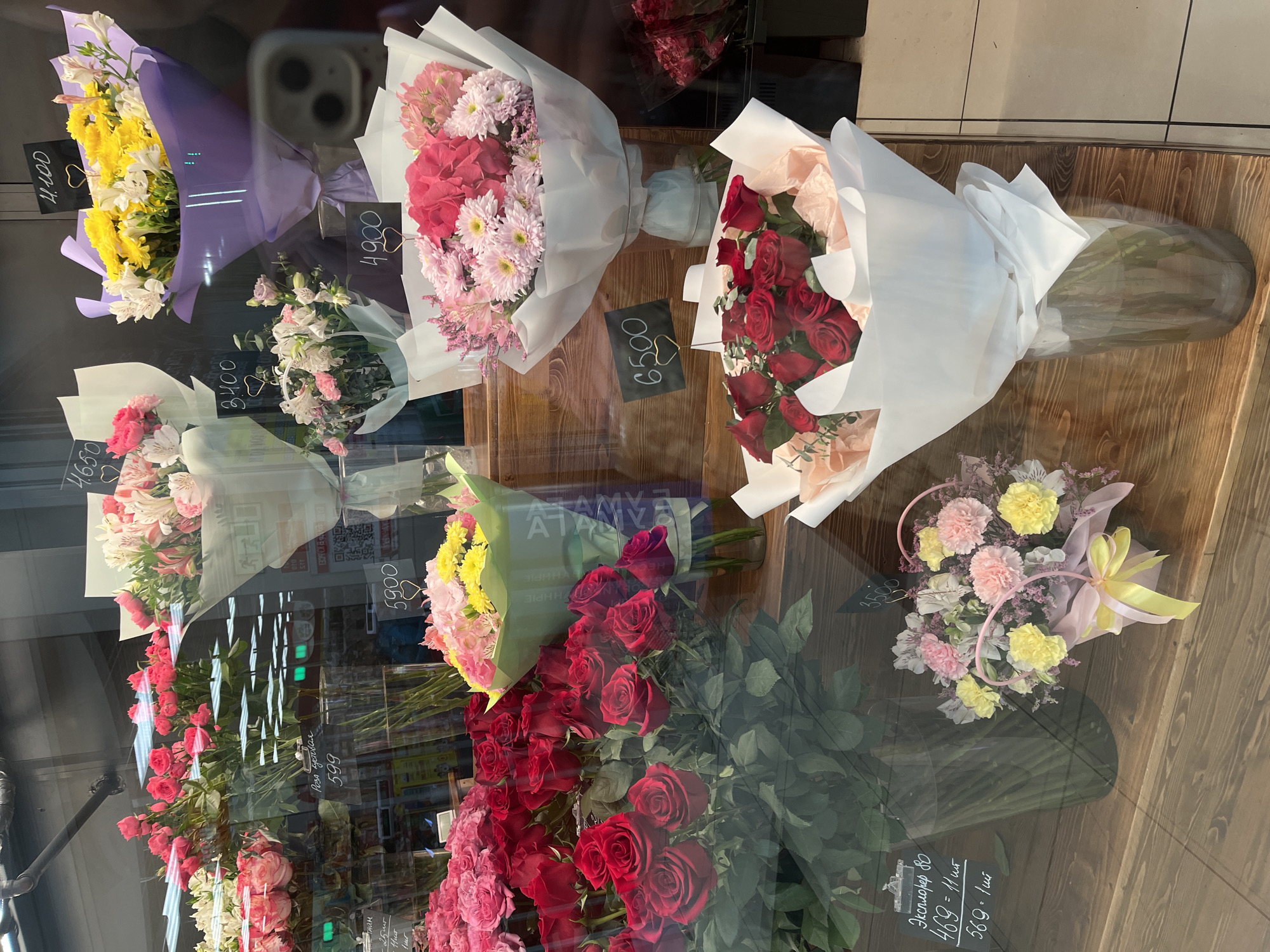 The image shows a flower shop display in Petropavlovsk-Kamchatsky, Russia. There are several wrapped bouquets of flowers, including roses, chrysanthemums, and alstroemeria. The bouquets have price tags displayed in front of them. Additional flowers, including roses, are in vases at the bottom of the display. The reflections on the glass show details of the street outside the shop. The display includes both single stem roses and bouquets of various flower types. The flowers are displayed on a wooden surface. Handwritten signs in Russian provide details about the flowers and prices.
