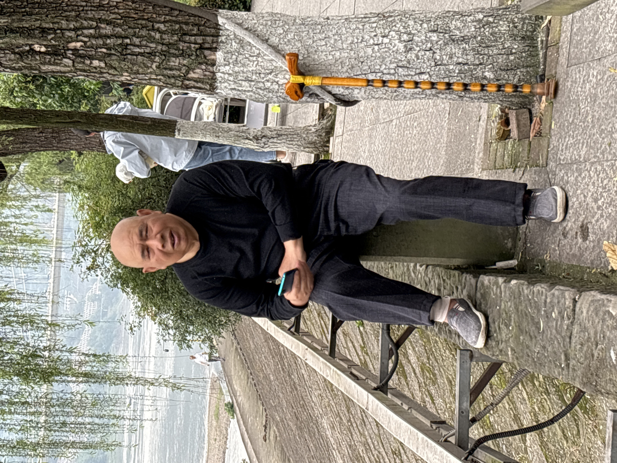 A man is sitting on a ledge. He is bald and wearing a black long-sleeved shirt and dark pants with grey shoes. He is holding a blue smartphone in his left hand. The ledge is grey and overlooks a body of water. A metal railing is situated between the ledge and the water. Weeping willow branches hang down from the upper left. In the background, another person can be seen walking along the waterfront. Behind the man is a tree with white-painted sections on the trunk. A wooden walking stick with a decorative handle rests against the tree. The location is Jiangjin, China.