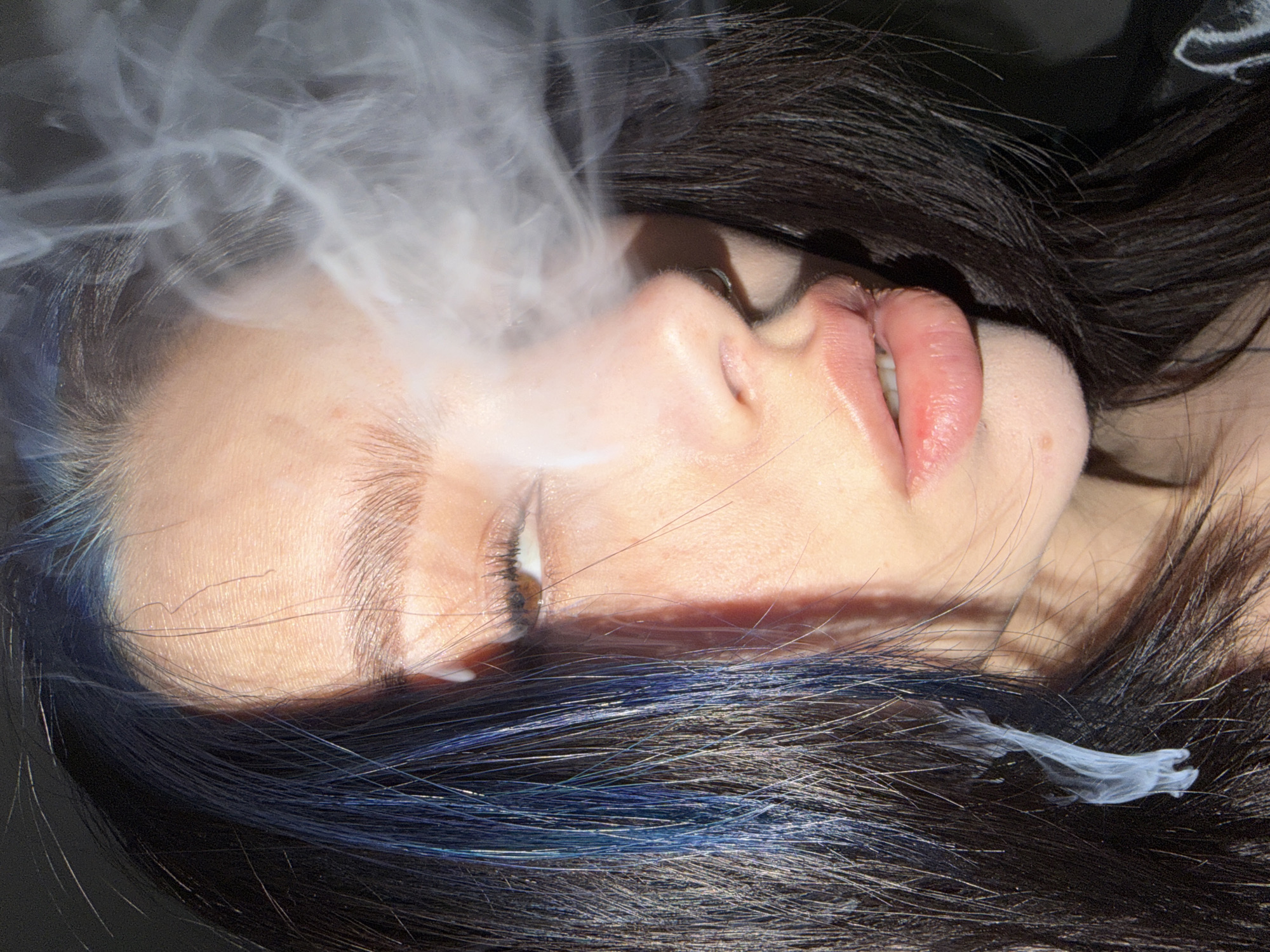 A close-up, high-angle shot captures a person, likely female, lying down with their head tilted, as smoke or vapor billows from their mouth. The individual's face occupies the central part of the frame, with eyes closed or mostly closed, and lips slightly parted. The skin appears fair, with visible pores and light texture. Dark hair, featuring distinct blue streaks, frames the face and extends across the bottom and top of the image, contributing to the dark background.
The primary activity is the exhalation of a significant cloud of white smoke or vapor, which originates from the person's mouth and spreads upwards and outwards, partially obscuring the forehead and hair. Smaller tendrils of smoke are also visible on the hair near the neck. The perspective is intimate, focusing solely on the face and hair, without any discernible background elements or location cues within the image itself. The provided context indicates the location is Khimki, Russia.
No specific emotions are strongly conveyed due to the closed eyes, but the scene suggests a moment of calm or introspection. There is no visible text in the image. The lighting highlights the skin and lips, contrasting with the dark hair and shadowed areas.