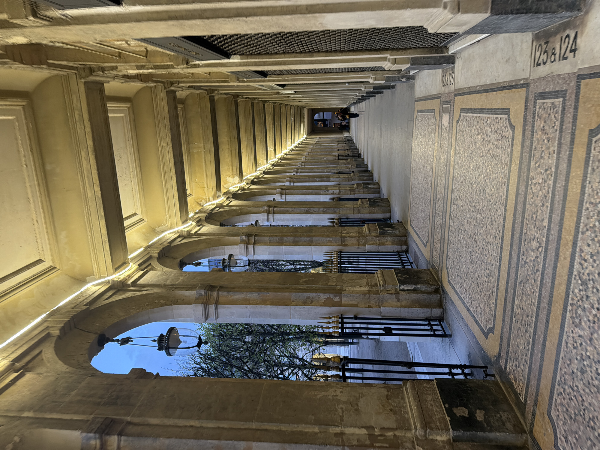 The image depicts a long, illuminated arched arcade within the Palais Royal gardens in Paris, France. The perspective is from within the arcade, looking down its considerable length.
The arcade is formed by a series of heavy, light-colored stone pillars supporting rounded arches. The underside of the arches and the ceiling are painted in a pale yellow, featuring prominent horizontal beams. Linear LED strip lighting is affixed to the top interior edge of the arcade, providing a consistent glow. Ornate, dark-framed lanterns with clear glass globes hang from the apex of multiple arches, spaced regularly along the corridor.
The floor is covered with a patterned mosaic of beige, brown, and grey tiles, delineated by solid yellow-beige and dark grey stripes. Adjacent to the tiled floor on the right, black stenciled numbers "123 & 124" and "122 & 121" are visible on the concrete surface. The right side of the arcade is lined with establishments, most of which are secured by dark, corrugated metal roll-down shutters.
On the left side, a black wrought-iron fence with decorative gold-tipped spikes separates the arcade from an outdoor area. Beyond this fence, bare deciduous trees with minimal green leaf buds are visible against a twilight-blue sky. In the far distance, a single individual, dressed in dark attire, walks away from the camera deeper into the arcade. A second, indistinct figure is partially visible further down on the right side. The scene is illuminated by artificial light within the arcade, contrasting with the ambient natural light from the evening sky.