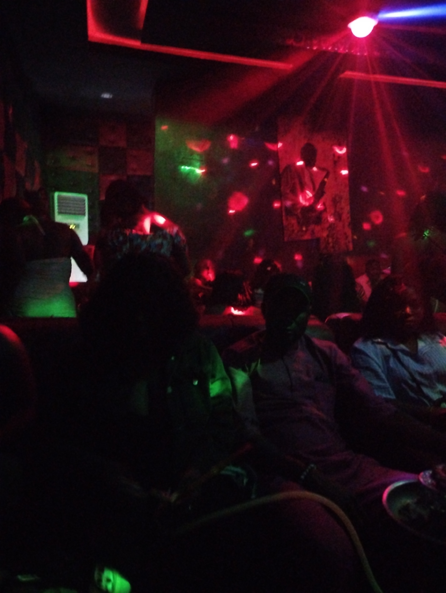 The image shows a dimly lit interior, likely a nightclub or lounge, in Gombe, Nigeria. Several people are seated and standing in the space, their faces and bodies partially obscured by the low lighting and shadows. Red and green lighting effects illuminate the scene, casting patterns on the walls and people. In the background, a painting or poster of a person is visible. In the foreground, individuals are seated, one wearing a light-colored long-sleeved shirt. A hookah is visible at the bottom of the frame. The atmosphere appears to be lively and social.