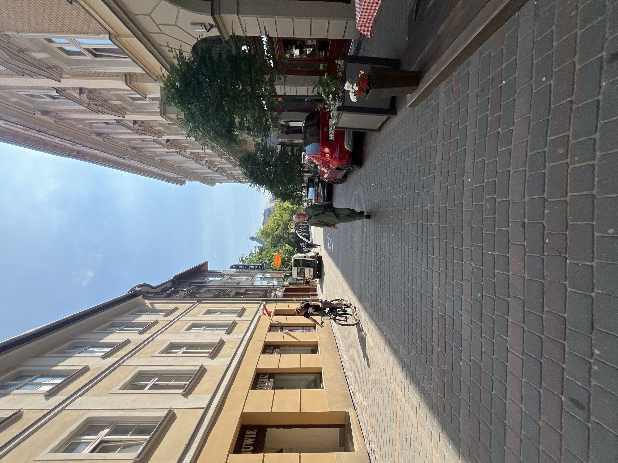 The image depicts a street scene in a city, with buildings lining both sides of a cobblestone street. The sun is shining, casting shadows on the street. On the left side of the street, there are two-story buildings with windows. A person on a bicycle is riding down the street. On the right side of the street, there are also buildings, some with outdoor seating areas. A red car is parked on the right side of the street, and a person is walking away from the camera. The sky is blue with some clouds. The street is relatively narrow, and there are trees and plants along the buildings.