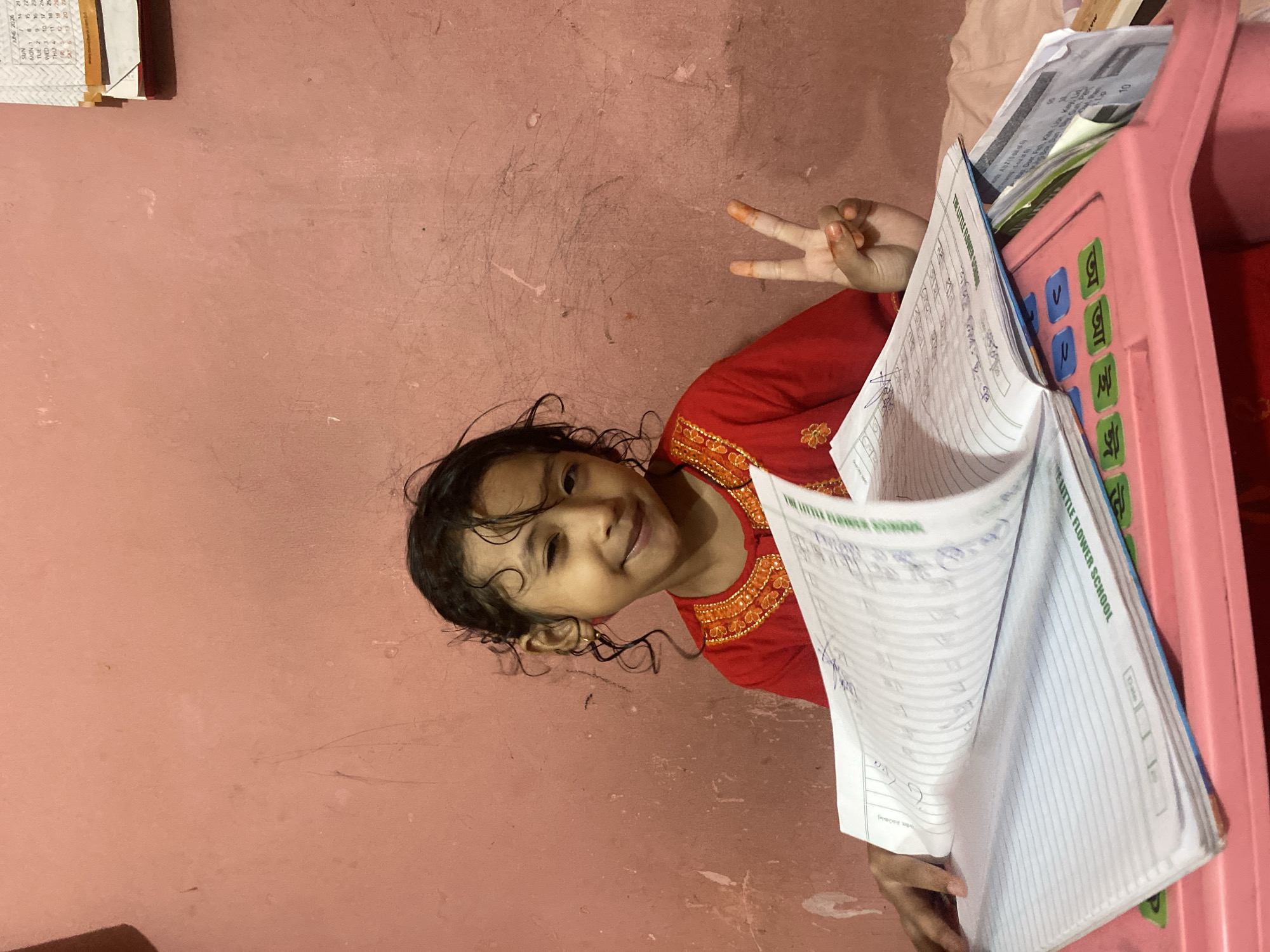 A young girl with curly dark hair, wearing a red and orange embroidered top, sits at a pink desk in front of a pink wall. She is smiling and making a peace sign with her right hand. A large open notebook, filled with lined paper and writing, rests on the desk in front of her. The notebook appears to be for "THE LITTLE FLOWER SCHOOL". A calendar from June 2024 is visible on the wall behind her. The scene is likely indoors, possibly in a home or classroom environment, in Dhaka, Bangladesh.