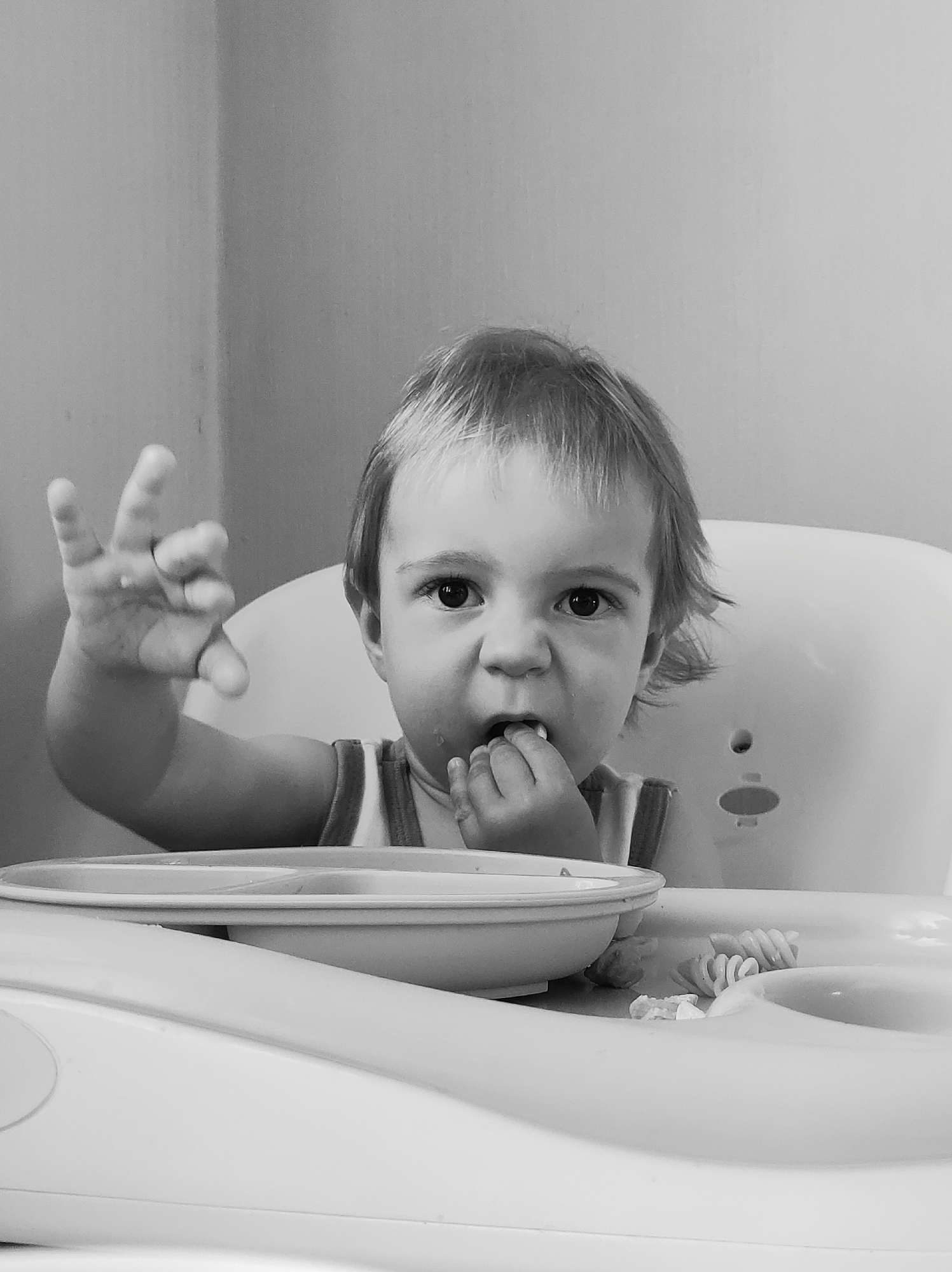 A monochrome, medium-shot image shows a young child in a high chair, engaged in eating. The child, with short, light hair, is seated behind a white high chair tray and bowl. Pasta pieces are scattered on the tray. The child is eating with their right hand near their mouth, and the left hand is raised with fingers splayed. The background is a plain, light-colored wall. There is no information to indicate that this image was taken in the city of Chaikovsky, Russia.