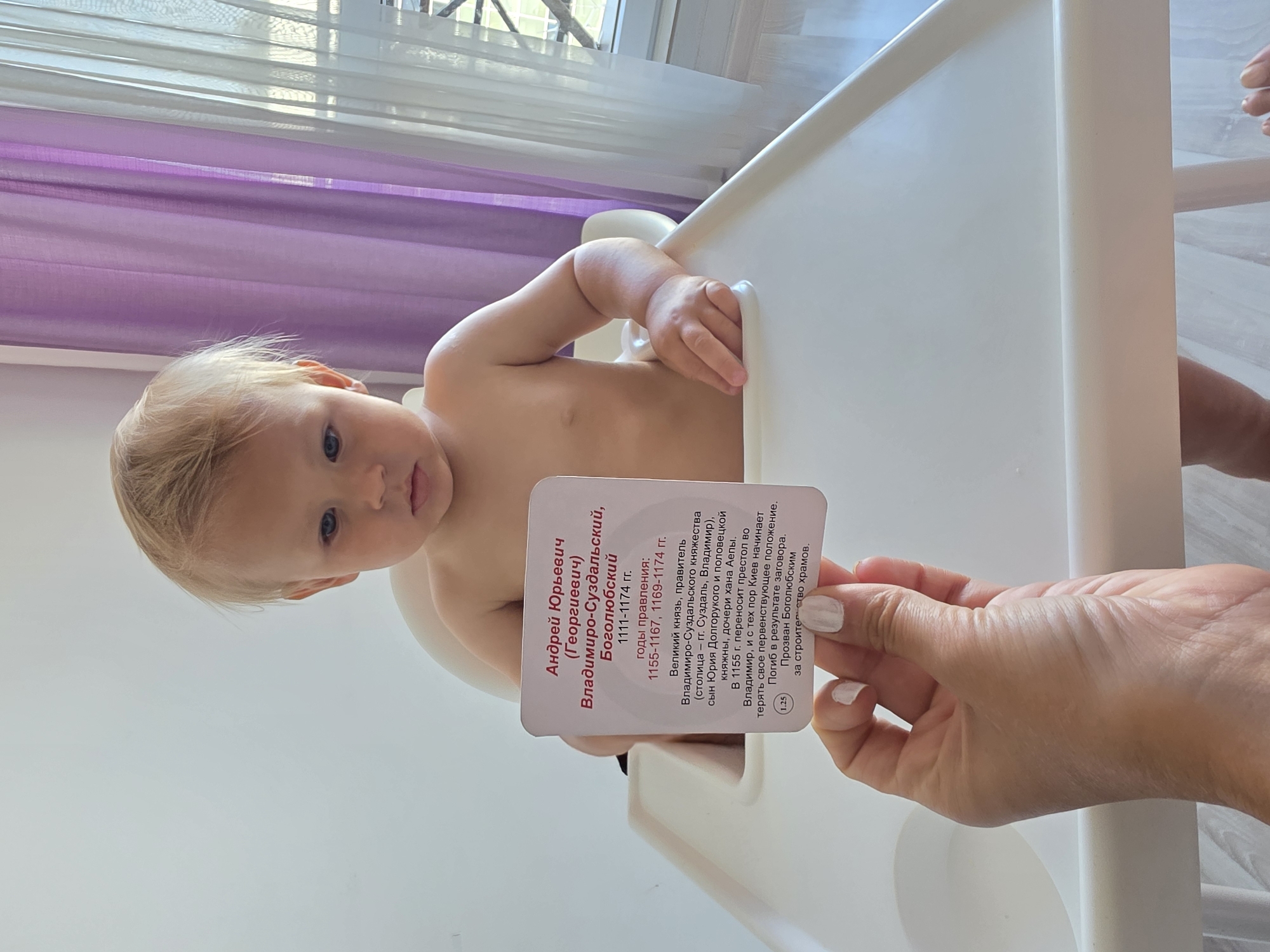 A fair-skinned, blond-haired toddler with blue eyes is seated in a white high chair, looking down with a neutral expression. The toddler is wearing a white paper card that reads "Андрей Юрьевич (Георгиевич) Владимиро-Суздальский, Боголюбский 1111-1174 гг." The card also contains biographical information in Russian about this historical figure, including his reign dates and titles. A woman's hand with white nail polish is holding the card up. The background consists of sheer purple and white curtains, and a window with a criss-cross pattern is visible behind them. The scene appears to be indoors, possibly in a residential setting, and there is no indication that this image was taken in Sochi, Russia.