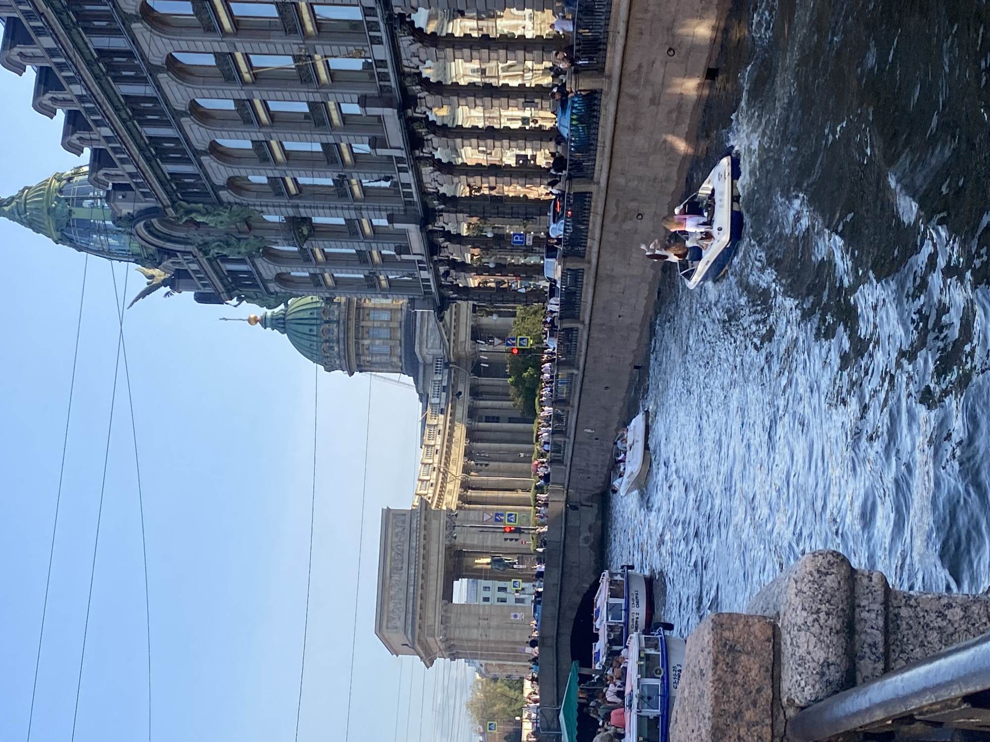 The image shows a canal in Saint Petersburg, Russia, with boats navigating the waterway. On the right, a large, ornate building with arched windows and a green domed roof is visible. This is likely the Singer House (Dom Knigi), also known as the House of Books, on Nevsky Prospekt. On the left, a grand colonnaded structure with a prominent archway is seen, which is part of the Kazan Cathedral. Several tour boats are on the canal, with passengers visible on deck. One smaller speedboat with four people is in the foreground, creating a wake. Pedestrians are seen walking along the embankment and on a bridge. The sky is clear and blue, indicating a sunny day.