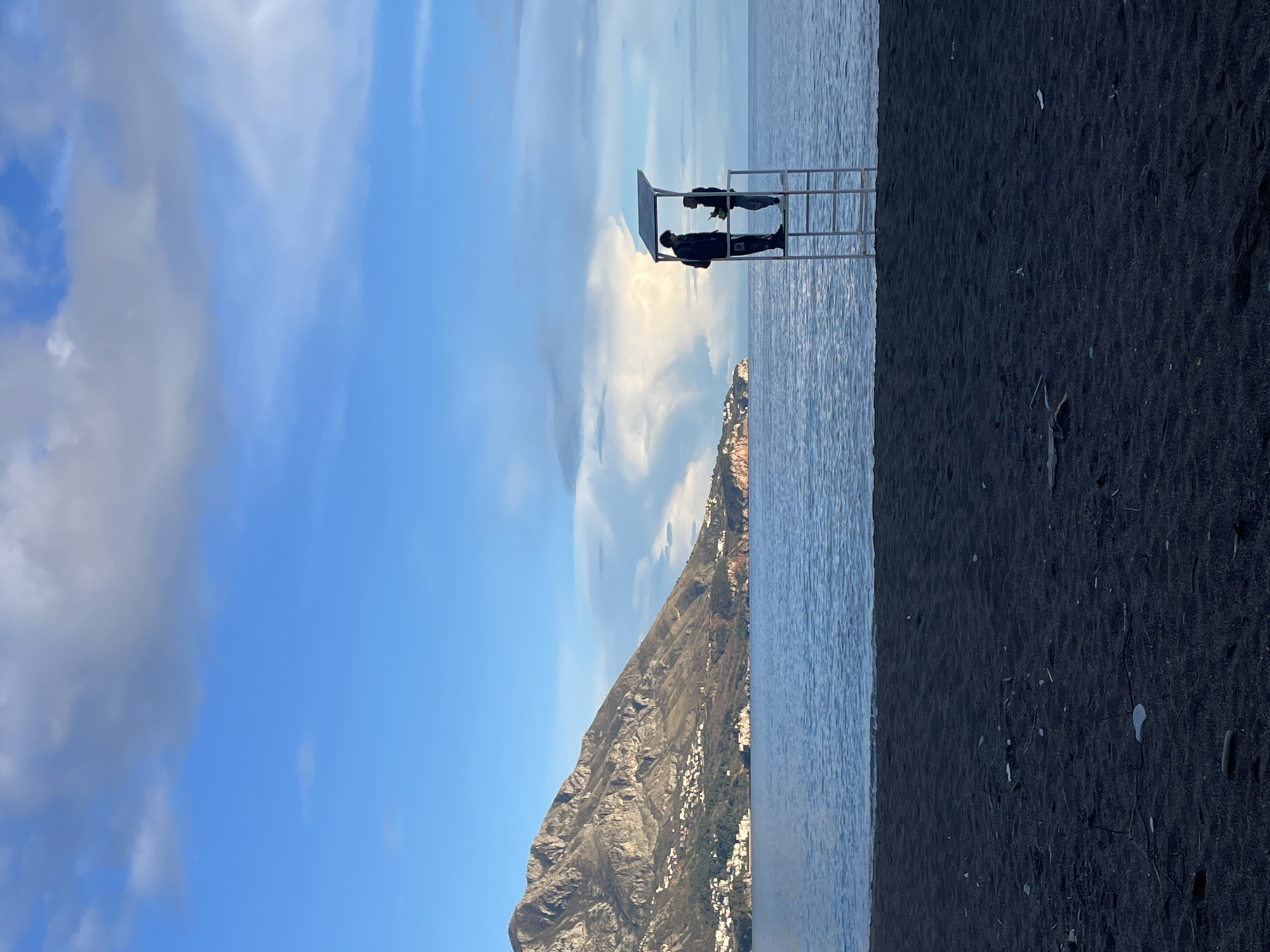 The image displays a coastal scene in Bečići, Montenegro, under a partly cloudy sky. The foreground consists of a dark grey/black sandy beach, scattered with small pieces of natural debris such as twigs and stones. The midground features calm, blue-grey sea water. Centered horizontally at the juncture of the beach and the sea, a white, metal multi-level lifeguard stand with a flat canopy is positioned. Two individuals are standing in silhouette on the top platform of this stand. The person on the left is dressed in what appears to be a jacket with a hood, carrying a backpack, and faces towards the right. The person on the right, in lighter clothing, faces towards the left, appearing to interact with the first individual, and holds a small, indistinct object in their right hand. In the background, a large, rugged mountain with visible rock formations and sparse vegetation dominates the left side of the horizon. Numerous white buildings are visible clustered along the base and lower slopes of this mountain, extending towards a distant headland. The sky above is bright blue, transitioning to areas with scattered grey and white clouds, including a prominent, brightly illuminated cumulus cloud formation over the sea in the middle-right.