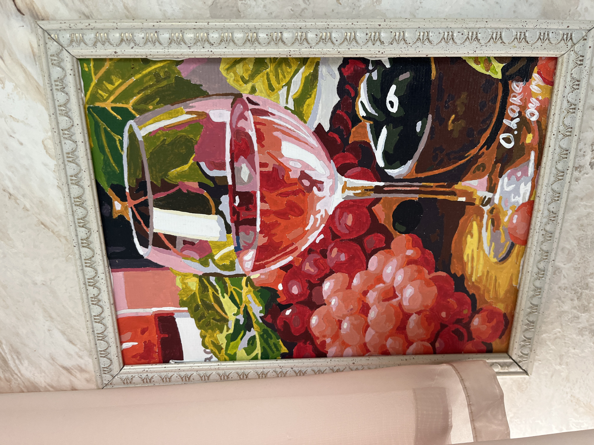 The image shows a framed painting of a glass of red wine and grapes. The painting is in a bright, stylized manner, with bold colors and distinct brushstrokes. The wine is depicted with a gradient of reds and pinks, reflecting light. A bunch of grapes, in varying shades of red and pink, is on the left side of the painting, and another dark cluster is on the right, possibly olives in a bowl. The background features abstract shapes and colors, including green leaves and a reddish-orange area. The signature "O. LORA 04.19" is visible in white paint at the bottom right of the painting. The painting is displayed against a textured, off-white wall, with a sheer pink curtain partially visible on the left side of the frame. There are no people or animals visible in the image. The painting appears to be a still life. The provided crops focus on the frame, the textured wall, and the details of the painting itself. There is no information within the image that suggests a location in Vladimirovka, Russia.