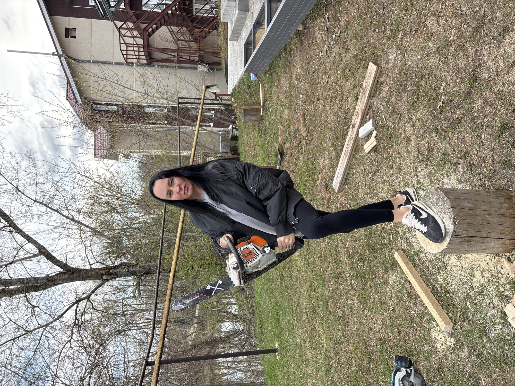 An outdoor scene, likely a backyard or semi-rural residential area in Chaikovsky, Russia, shows a woman posing with a chainsaw. She stands centrally, looking directly at the camera with a neutral expression. She has long, dark hair and wears a black bomber jacket over a grey t-shirt, black leggings, and black Nike sneakers.
Her right foot is resting on a large, freshly cut wooden log or tree stump, surrounded by sawdust. She holds an orange and white Stihl MS 181 chainsaw. The chainsaw's blade features a graphic of crossed axes and the "STIHL" logo. Nearby on the grassy ground are several wooden planks and a blue and black circular saw.
In the mid-ground, a rusty metal fence or framework stretches across the scene. The background reveals residential buildings with reddish roofs and light-colored walls, one featuring an external metal staircase. Further to the right, a concrete structure with metal beams appears to be under construction. Trees with sparse, early spring foliage are visible against an overcast sky, suggesting daytime on a cloudy day. The scene depicts activity related to wood cutting or construction.