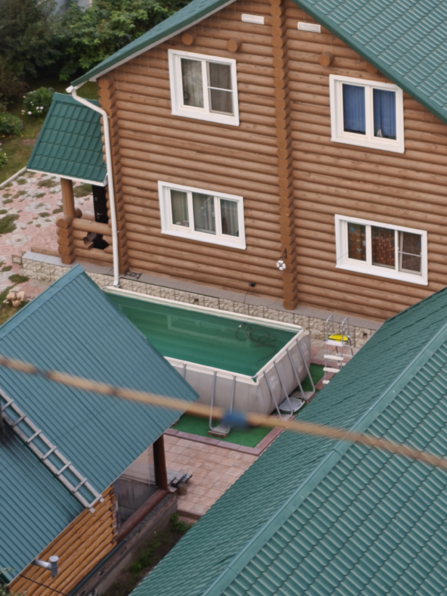 The image shows an aerial view of a house with a green metal roof, surrounded by greenery and a swimming pool. The house is constructed of wooden logs and has several windows with white frames. A swimming pool with green water is visible in the foreground, next to a ladder. The scene appears to be a residential area in Rubtsovsk, Russia, based on the provided context.