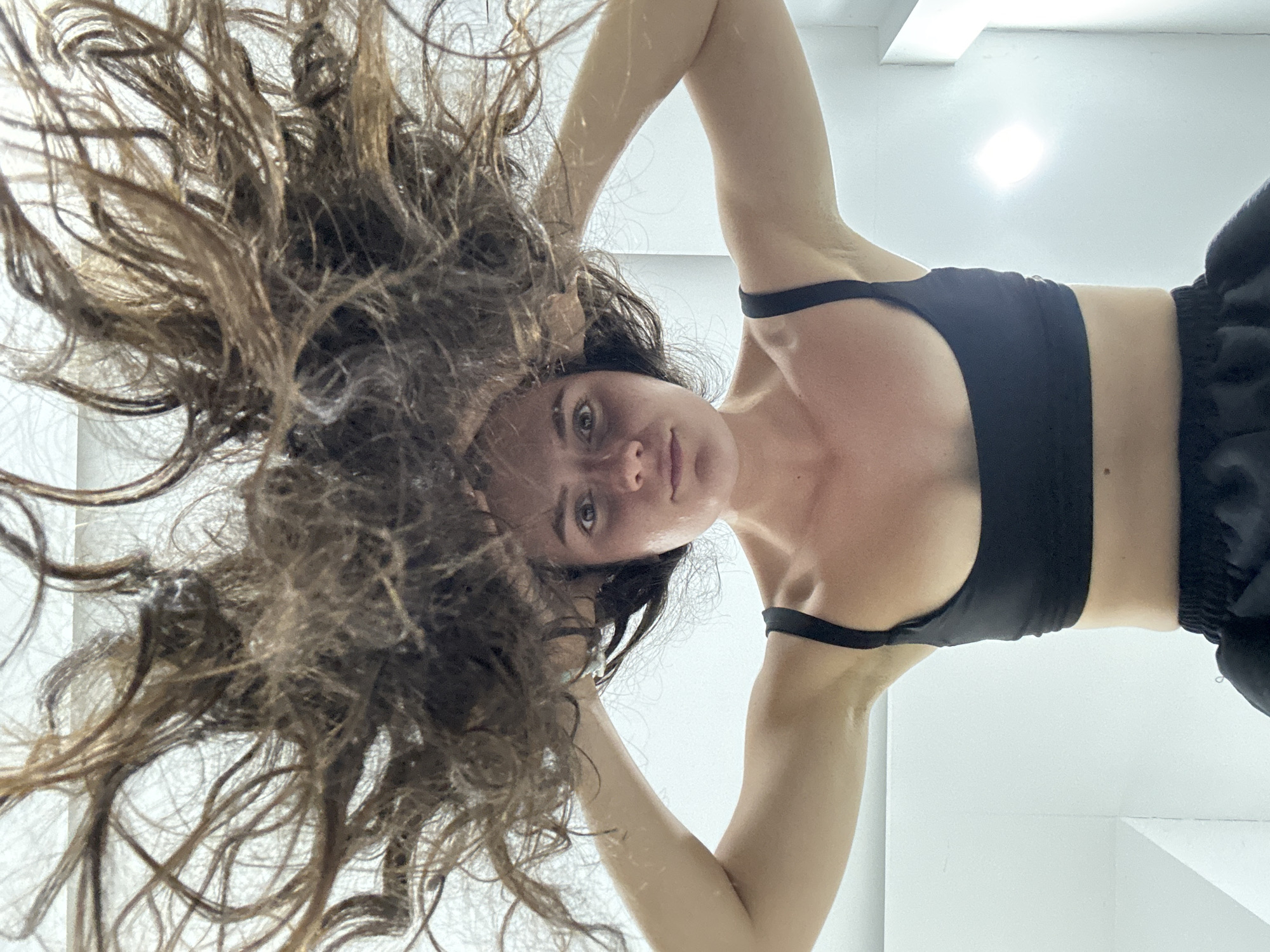 A young woman lies on her back in an indoor space with bright white walls and ceiling. She is wearing a black sports bra and dark athletic pants. Her arms are raised above her head, exposing her torso. Long, messy, brown hair surrounds her head and spreads out across the floor. The setting may be a gym or exercise studio, given the athletic attire and the stark, uncluttered background. The location in Mũi Né, Vietnam is not discernible from the visual information provided.