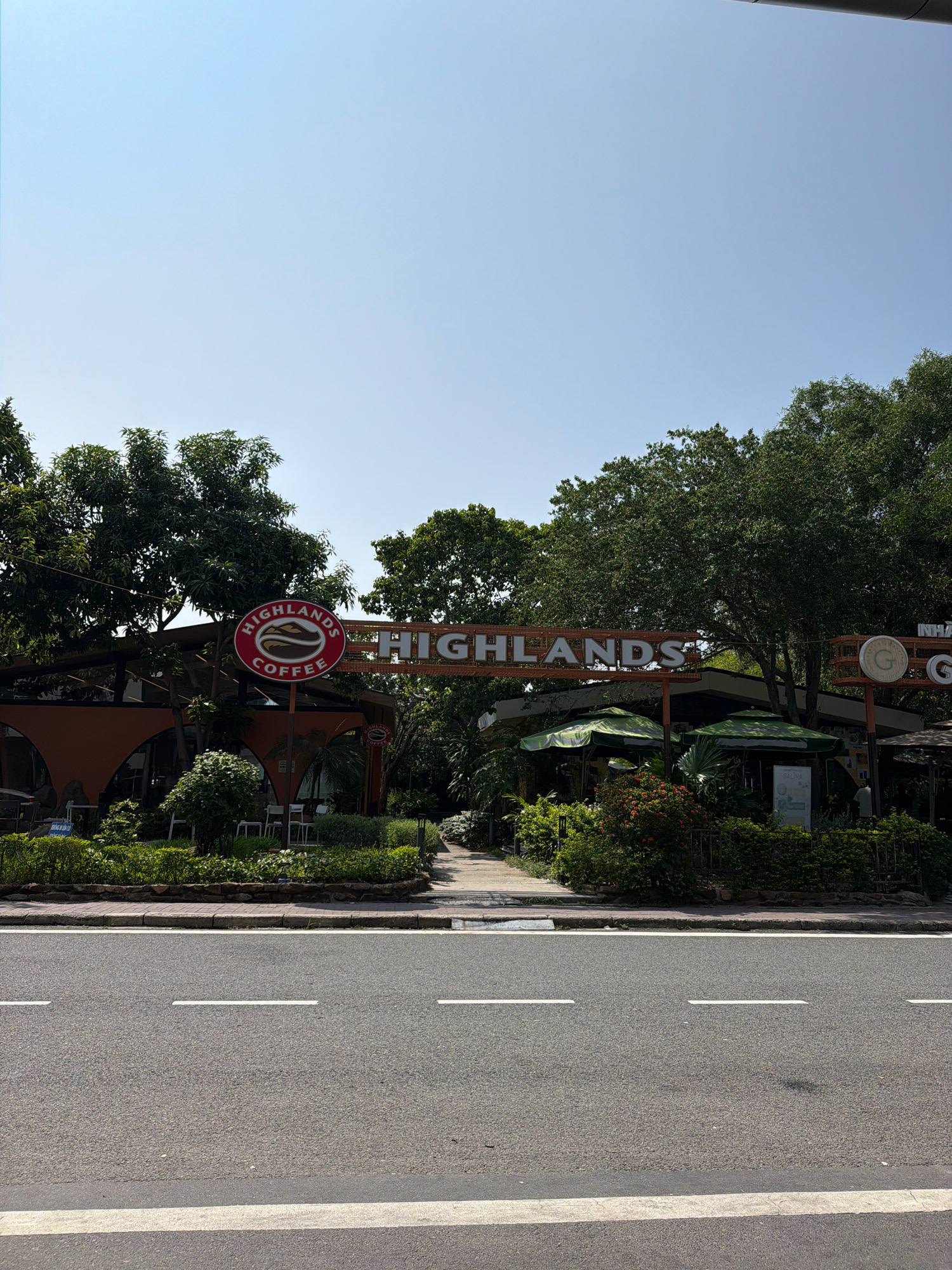 The image captures a scene outside a Highlands Coffee establishment in the city of Cam Ranh, Vietnam. The foreground features a section of asphalt road with white lane markings. Beyond the road, a sidewalk separates it from a landscaped area with bushes and a stone border.
Dominating the midground is the coffee shop, characterized by its orange arched facade and large windows. A prominent sign with "HIGHLANDS COFFEE" in white lettering against an orange background is visible. To the left, a circular logo for Highlands Coffee, featuring a coffee bean graphic, is mounted on a pole. Several green umbrellas provide shade over outdoor seating areas. Lush green trees and foliage surround the building, suggesting a tropical or subtropical environment. The sky above is clear and blue.
In the background, additional trees and part of another building are visible. A person can be seen standing near the right side of the Highlands Coffee building. The lighting suggests it is daytime, with the sun casting shadows.