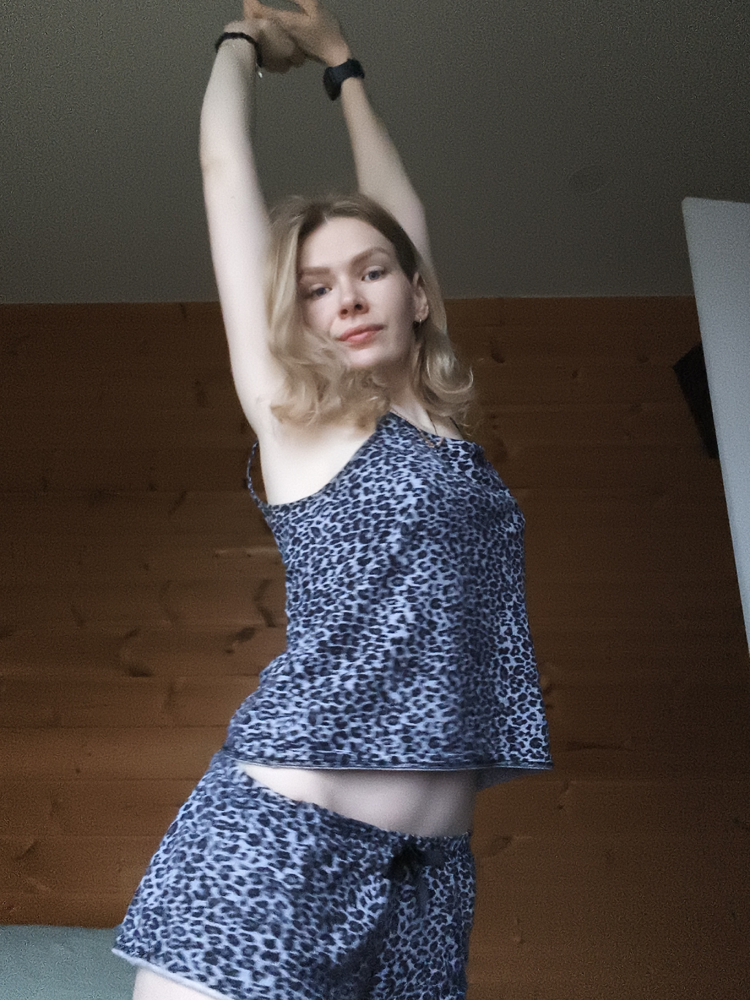 A fair-skinned woman with shoulder-length blonde hair stands indoors, wearing a matching blue and grey leopard-print tank top and shorts. She has both arms raised above her head, slightly bent at the elbows, with a dark-colored band on each wrist. The background consists of a wood-paneled wall and a white doorframe on the right. The location is in the city of Русский Вожой, Russia.