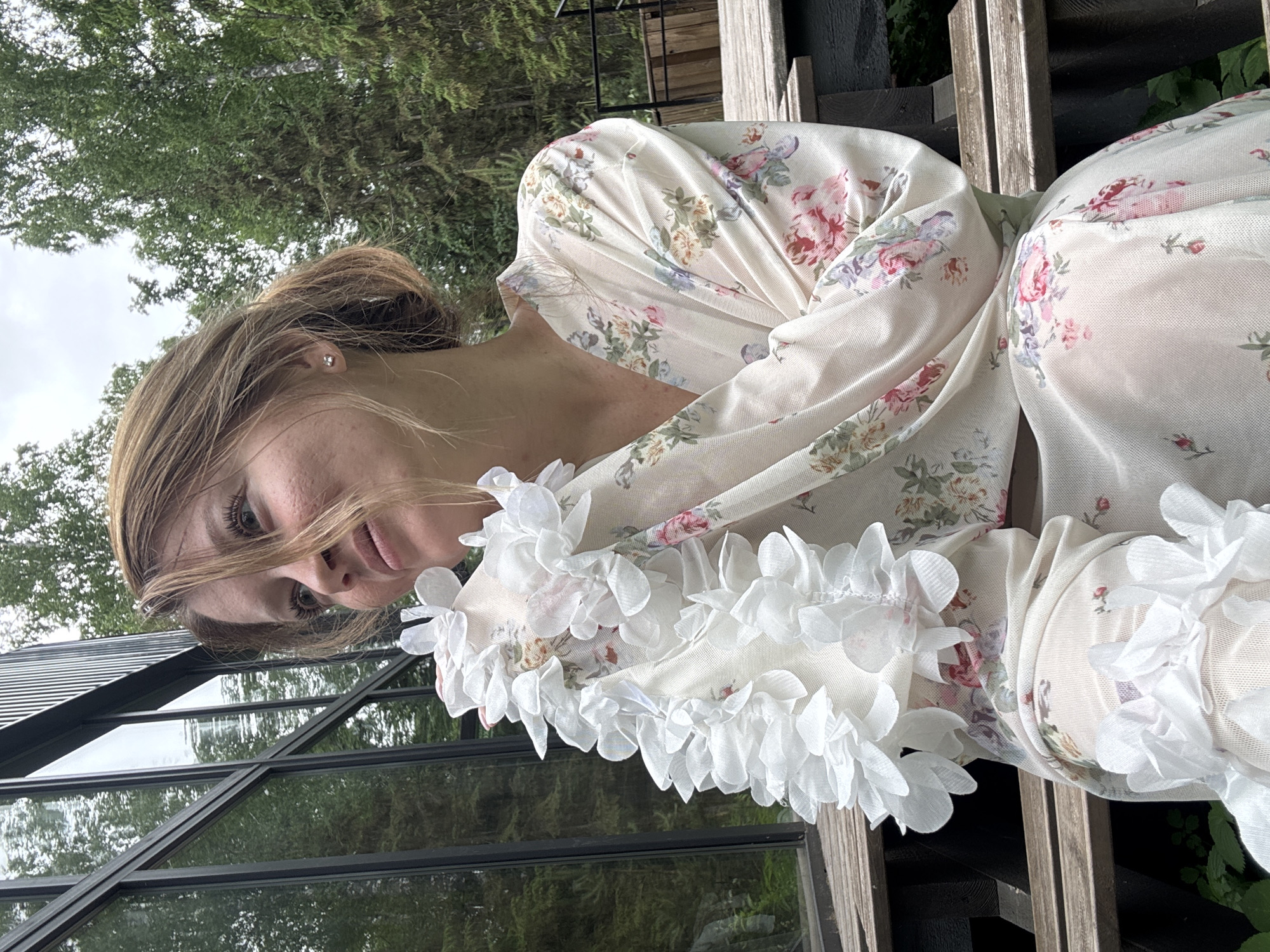 A young woman with light brown hair pulled back sits on a wooden bench outdoors, with a modern building with large windows to her left. She is wearing a light-colored robe with a floral pattern and white ruffled trim around the neckline. The background consists of green trees and an overcast sky. The image appears to be taken in a natural setting, possibly a yard or garden area associated with the building. No specific details about the location within Bakhtiyary, Russia are discernible from the image.