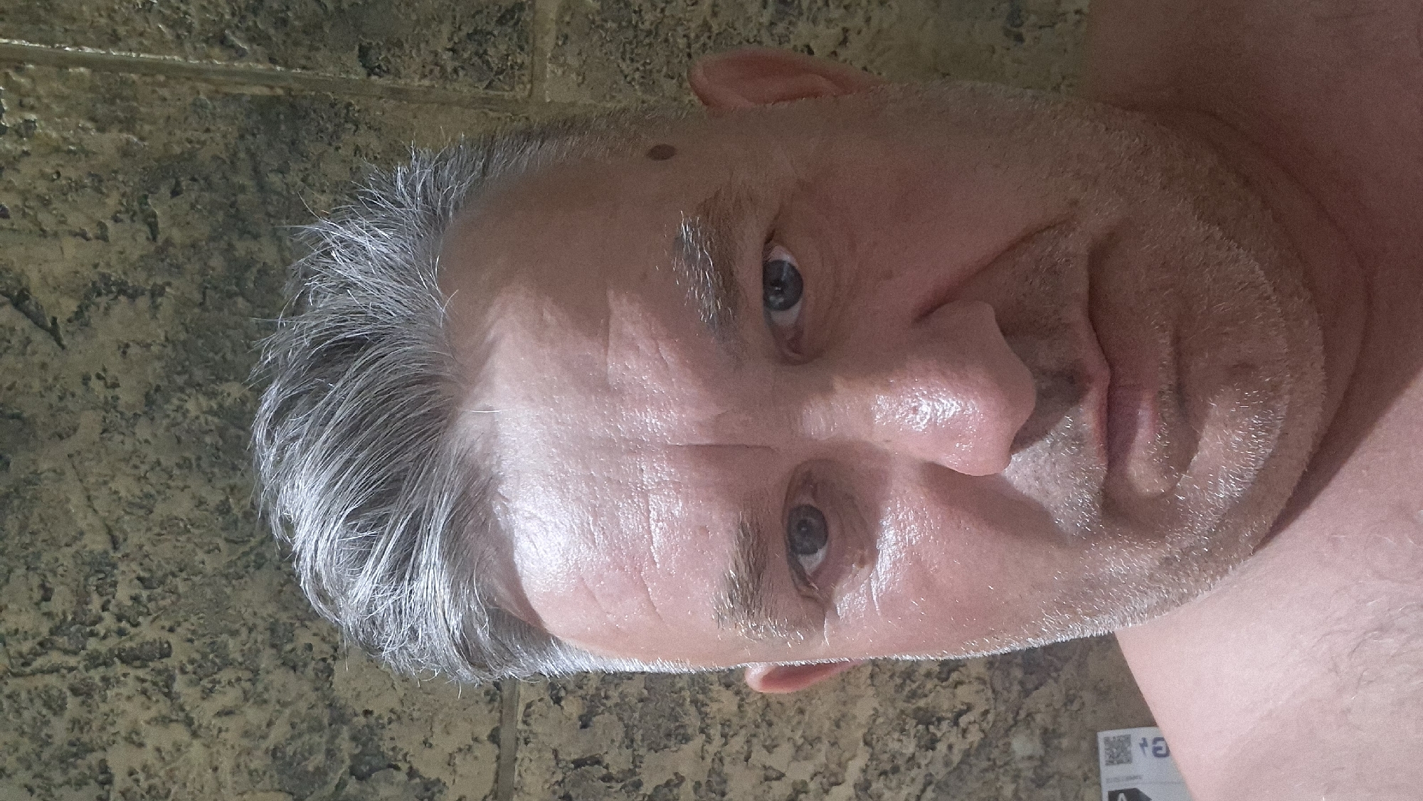The image shows a close-up selfie of a man with graying hair and a light beard. He is looking directly at the camera with a neutral expression. The background appears to be a textured wall. There are no other people or objects of note in the image. The location information provided is not verifiable from the image itself.