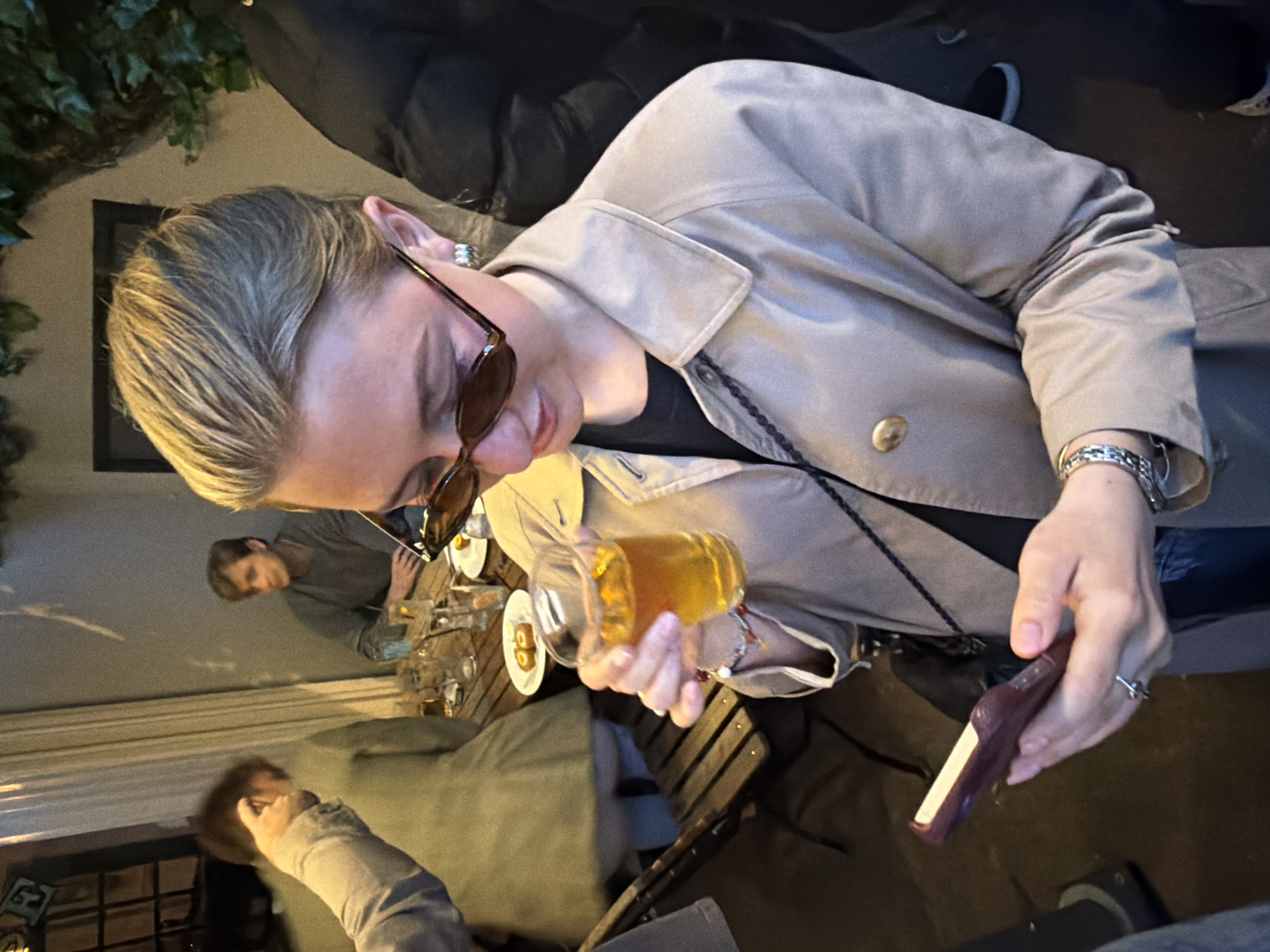 A woman wearing sunglasses and a beige jacket is holding a glass of amber liquid in her left hand and a small brown object in her right. She is sitting at a wooden table in an outdoor setting, presumably a bar or restaurant, with other people partially visible around the table. The location is possibly in London, United Kingdom, but the specific details of the environment are insufficient to confirm this precisely. There are other glasses and plates on the table. The woman is looking down at the objects in her hands.