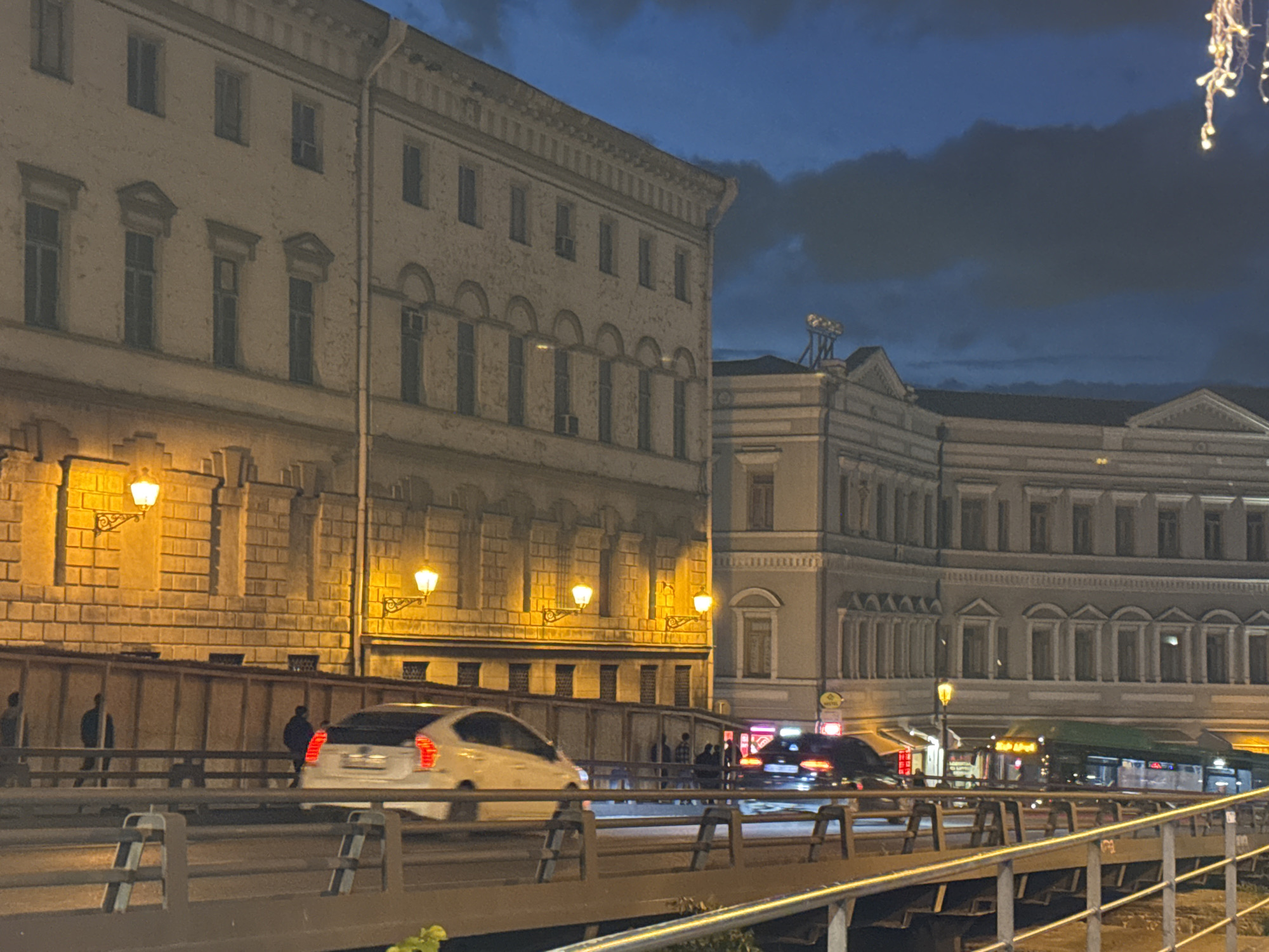 The image shows a street scene in Tbilisi, Georgia, at night. Two large, light-colored buildings with multiple windows form the backdrop. The buildings are illuminated with warm-toned street lamps. Pedestrians walk along a walkway between the building and the road. A white car drives on the road, partially obscured by motion blur. Further down the road, a dark car and a green bus are visible. The scene is viewed from a bridge, with the bridge's railing in the foreground. The sky is dark with clouds.