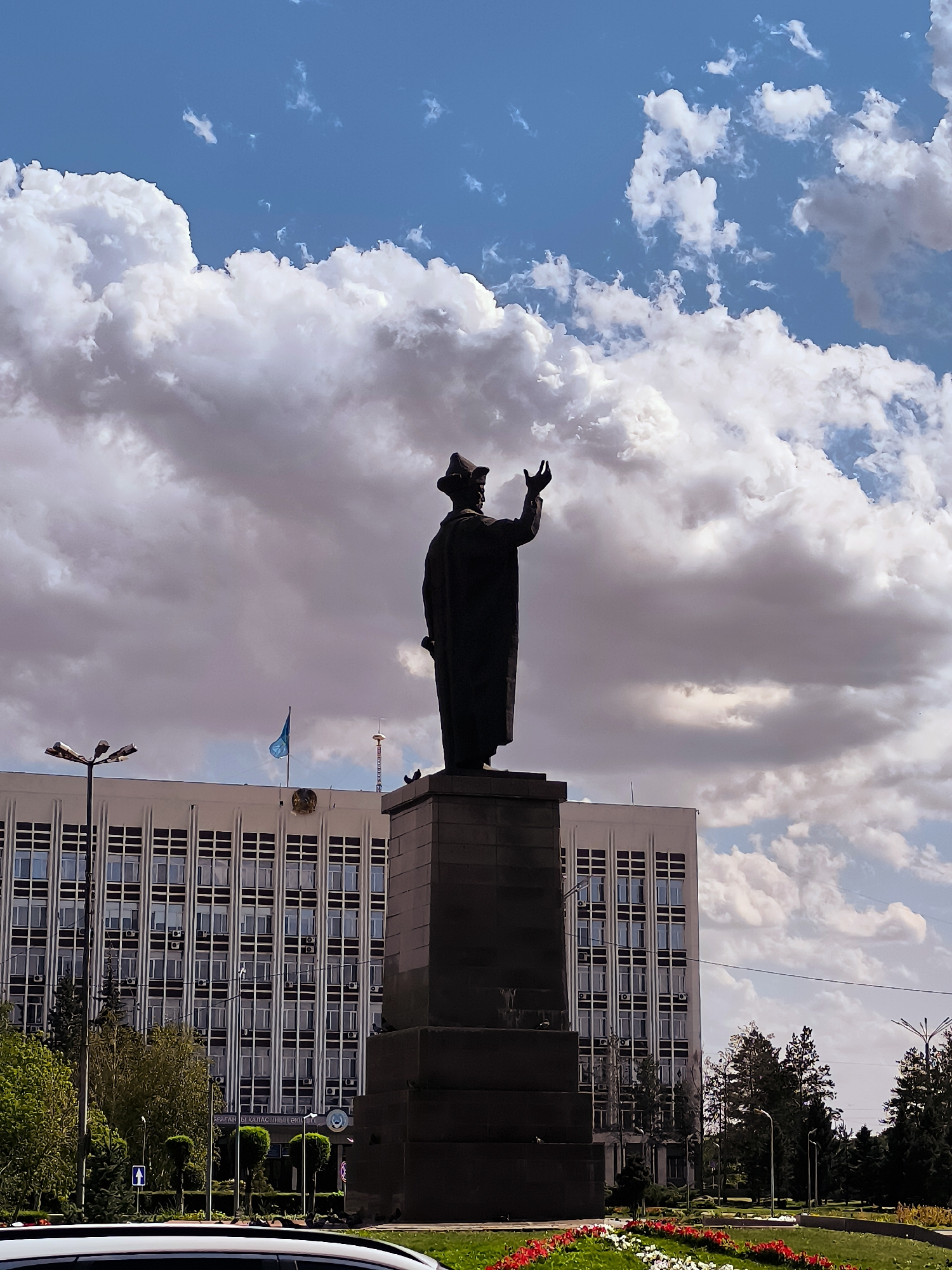 A statue of Abay Qunanbaiuly, a prominent Kazakh poet and philosopher, stands in a central square in Karaganda, Kazakhstan, against a backdrop of a large administrative building and a dramatic sky filled with white and gray clouds. The statue, made of dark material, depicts Abay in traditional attire, with his right arm raised. The building behind the statue is a multi-story structure with a repetitive pattern of windows and is adorned with a national emblem and a flag. The square features manicured trees, flower beds with red and white flowers, and streetlights. A car is partially visible in the foreground.