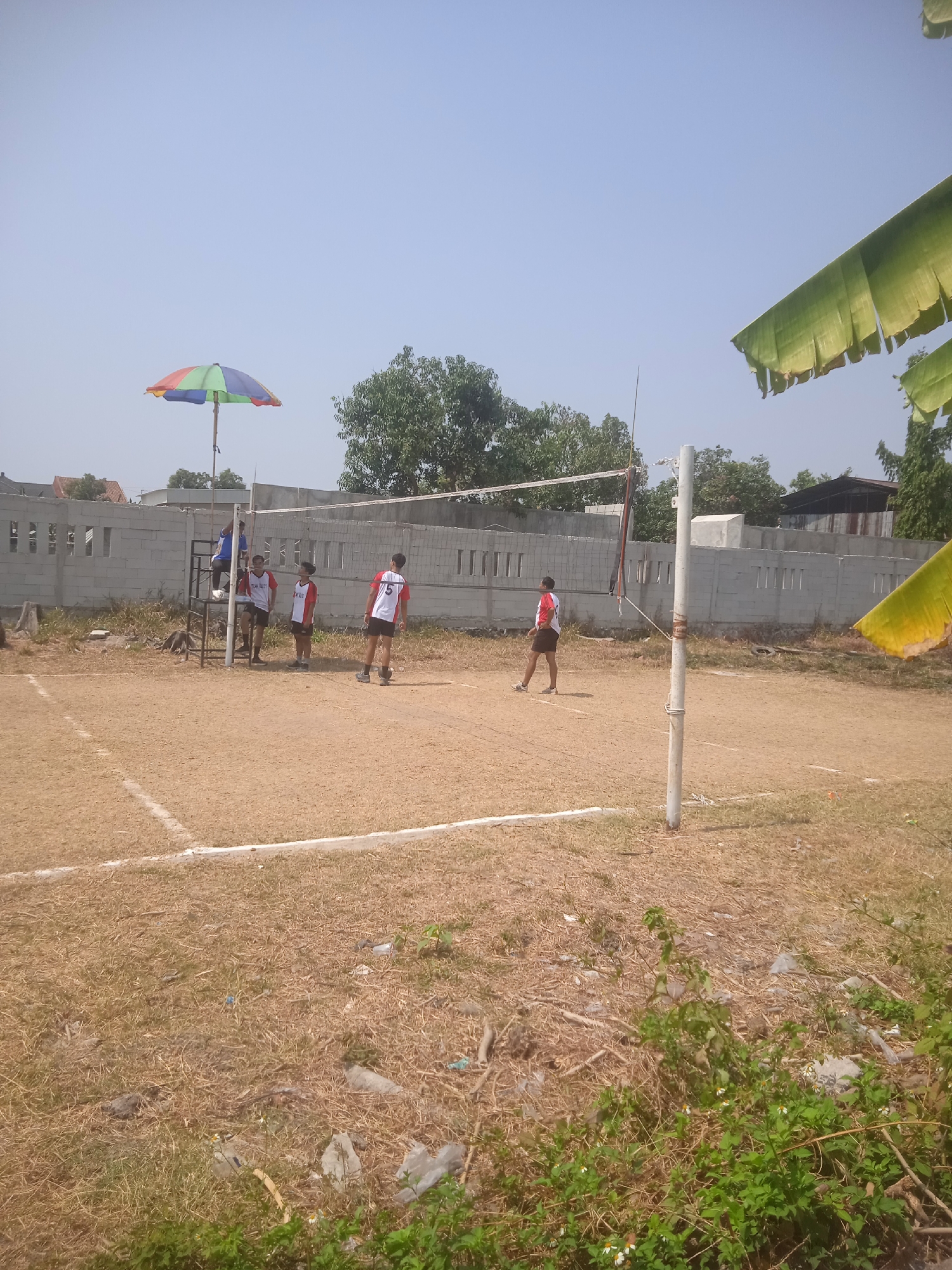 The image depicts a sandy volleyball court in Klodran, Indonesia, under a clear sky. A volleyball net is set up across the court, supported by white poles. Several young males in sportswear are present on and around the court. Two individuals are on one side of the net, one standing near the net and another on a referee's stand. On the other side of the net, two individuals are visible, one in the foreground walking and another in the background standing. A multi-colored umbrella provides shade near one of the net poles. The background shows a low, light-colored brick wall with vertical openings, beyond which are trees and some buildings. The ground of the court is predominantly dry and sandy, with some patches of sparse, dry grass and scattered debris. Banana leaves are visible hanging from the top right corner of the image.