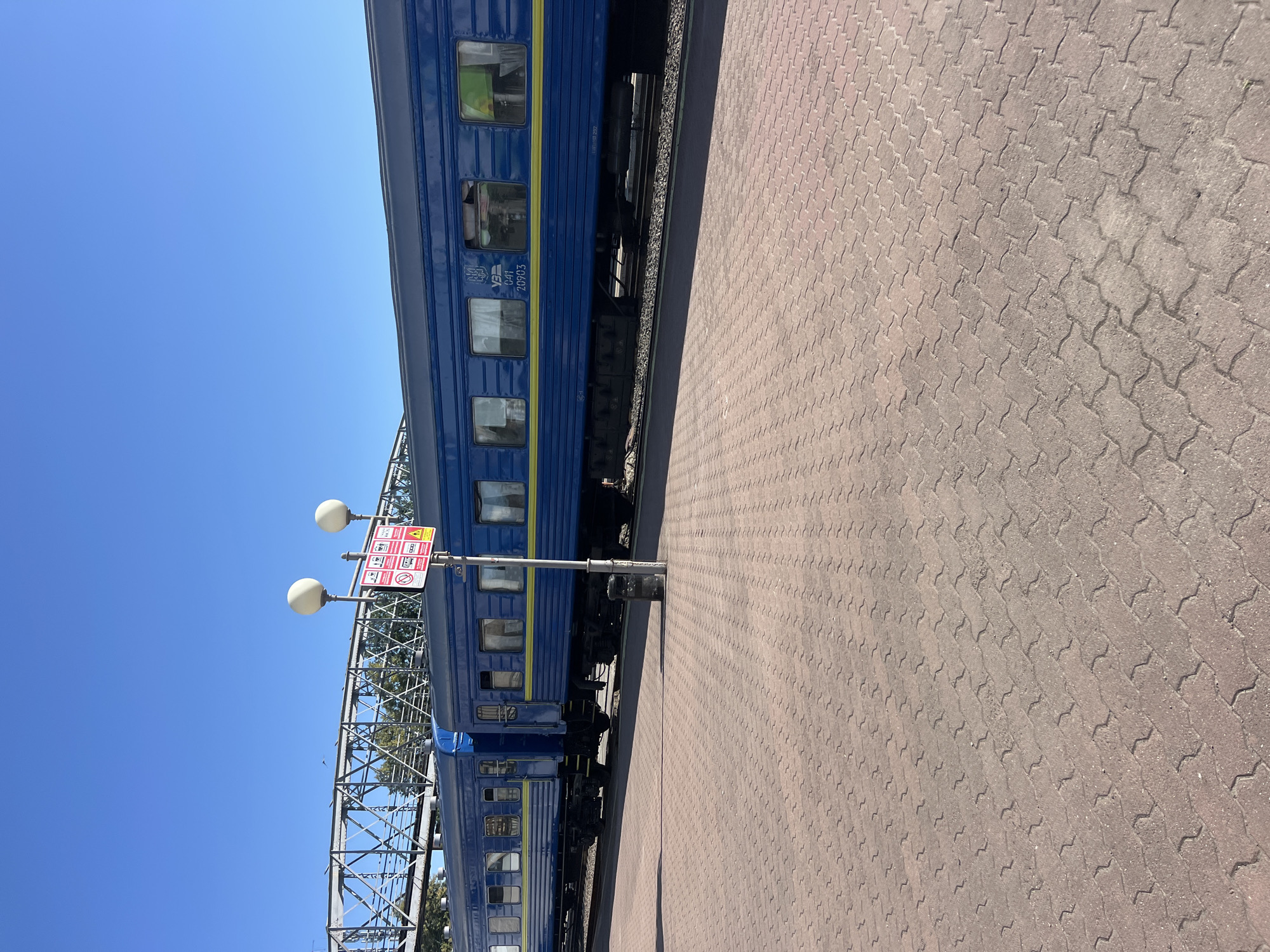 The image shows a blue passenger train at a station in Ivano-Frankivsk, Ukraine. The train cars have yellow stripes and visible windows. A paved platform with interlocking brickwork extends in the foreground, and a metal pedestrian bridge can be seen in the background. A signpost with multiple warning symbols and two streetlights are located on the platform near the train. The sky is clear and blue.