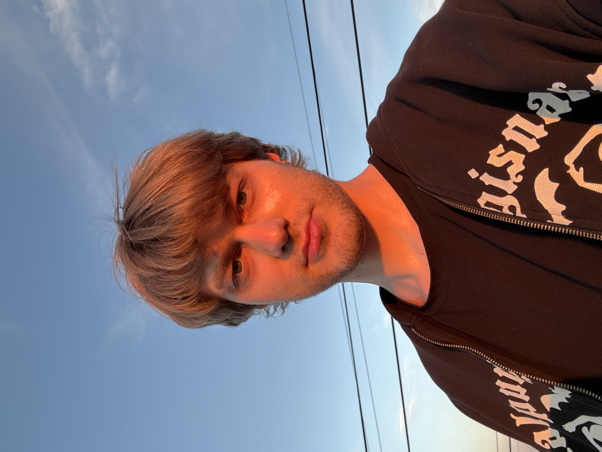 A young man with tousled brown hair is captured in a selfie against a clear blue sky. The setting sun casts a warm, orange glow on his face and hair. He is wearing a black t-shirt underneath an unzipped black hoodie with a white graphic design. The image appears to have been taken outdoors, with power lines visible in the background, suggesting an urban or suburban environment.